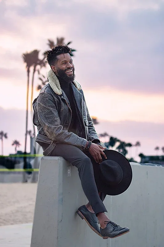 A man with a beard and dreadlocks sitting on a concrete ledge outdoors during sunset, holding a black hat, wearing a denim jacket with a shearling collar, dark pants, and black shoes, with palm trees in the background.