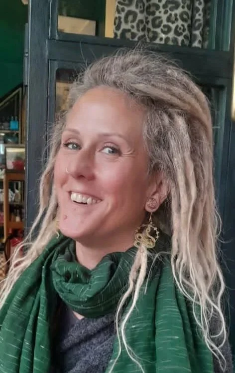 A woman with long, blonde, dreadlocked hair smiling, wearing a green scarf and earrings, standing inside a room with various objects in the background.