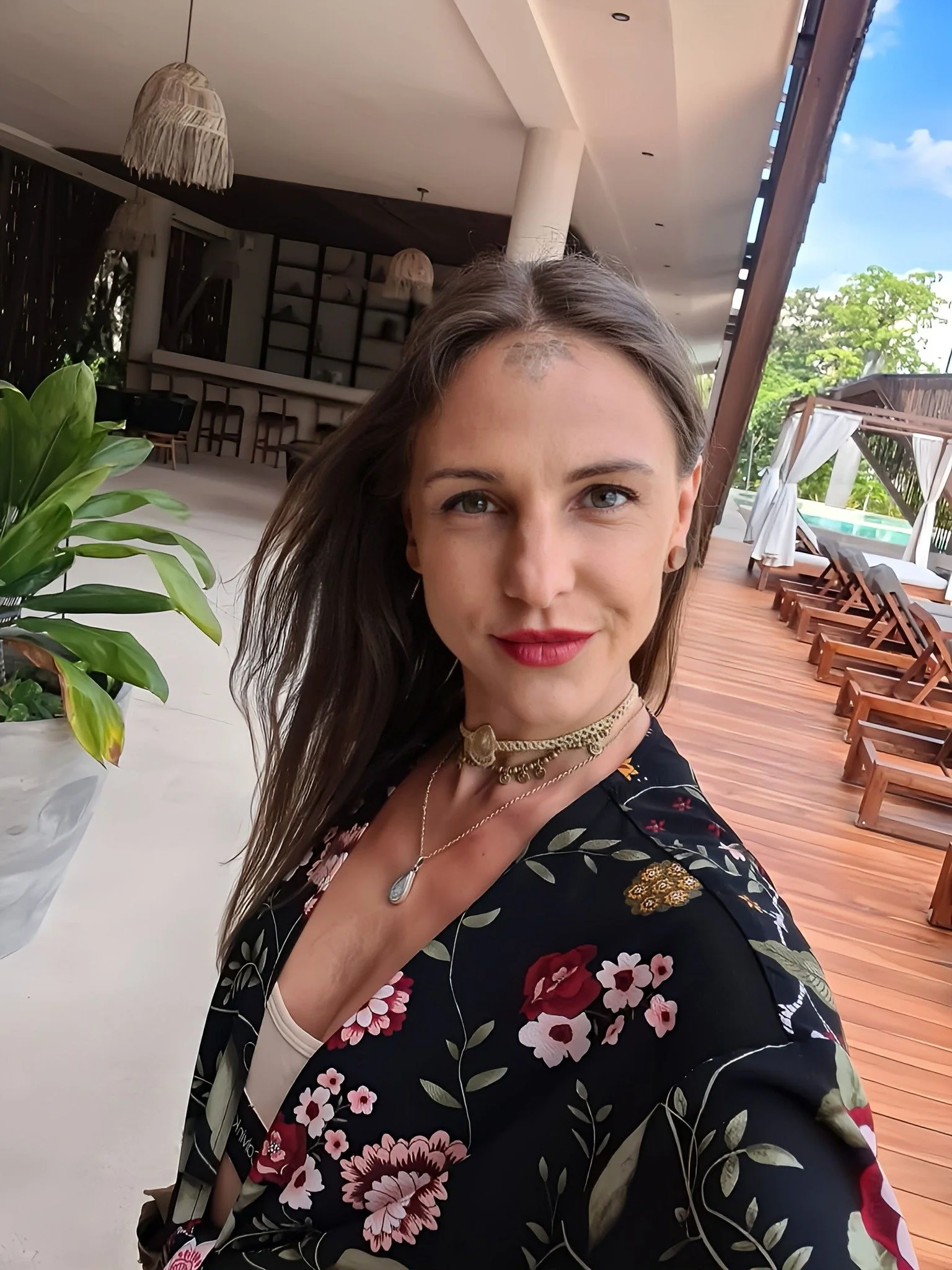 A woman with long brown hair, wearing a floral dress and jewelry, taking a selfie in a bright outdoor area with wooden deck chairs, greenery, and a pool.