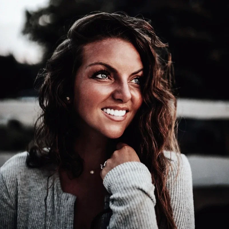 A smiling woman with wavy brown hair, wearing a gray sweater and a small necklace, looking upward and slightly to the side outdoors.