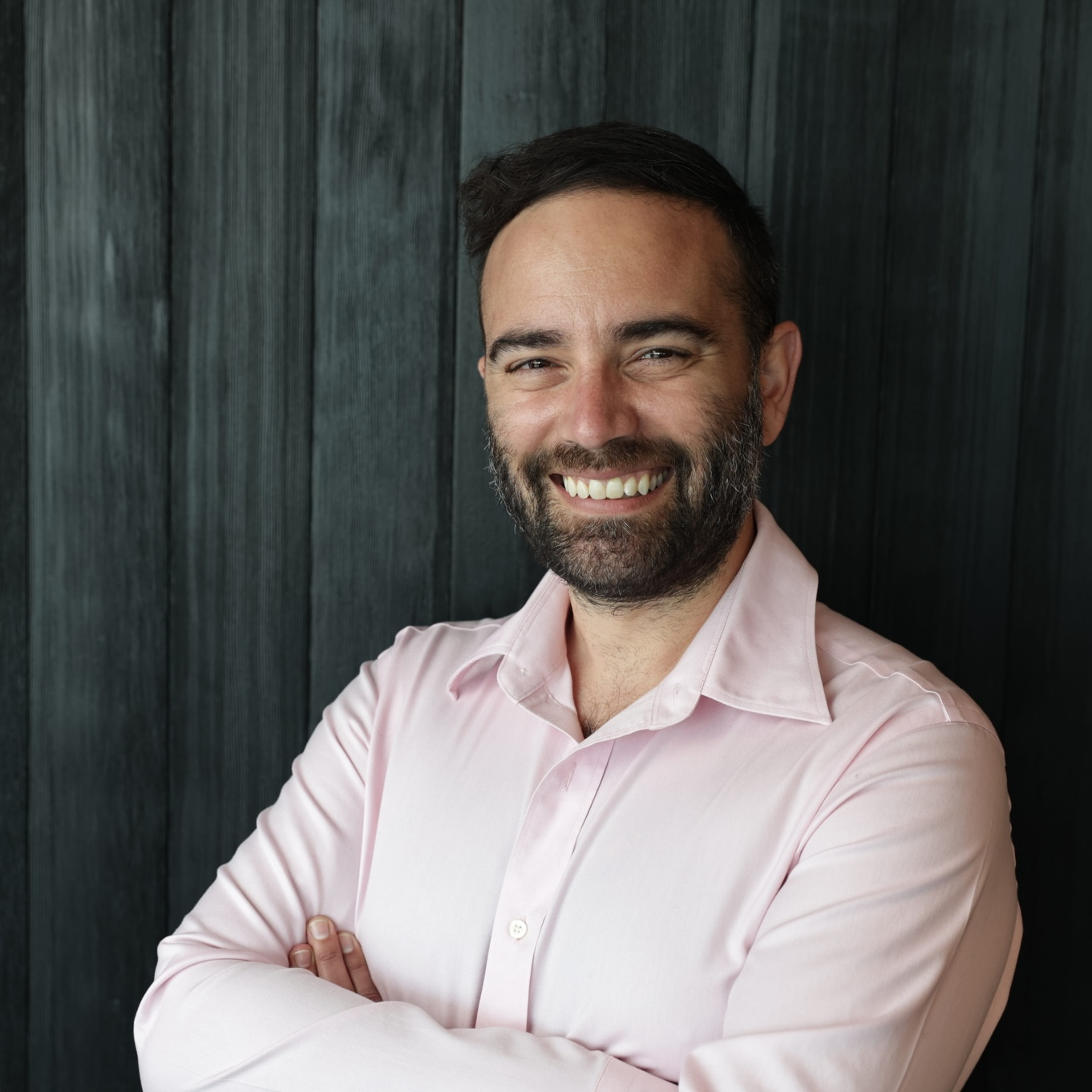 A smiling man with a beard and dark hair, wearing a light pink button-up shirt, standing against a dark wooden wall with arms crossed.