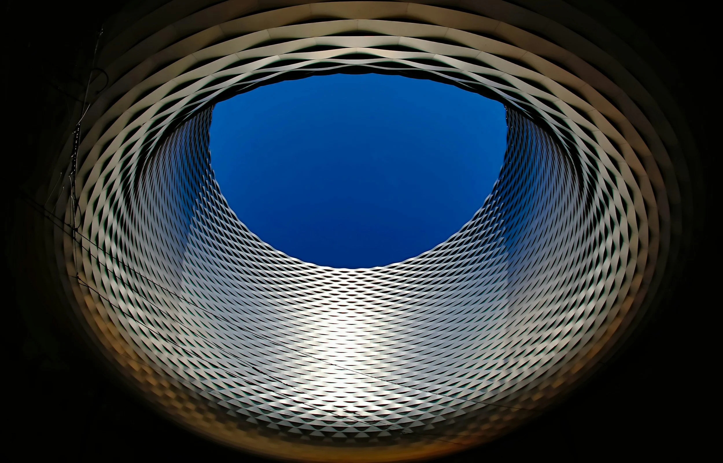 View looking up through a modern, oval-shaped building with a honeycomb-like exterior pattern, revealing a blue sky through the roof opening.