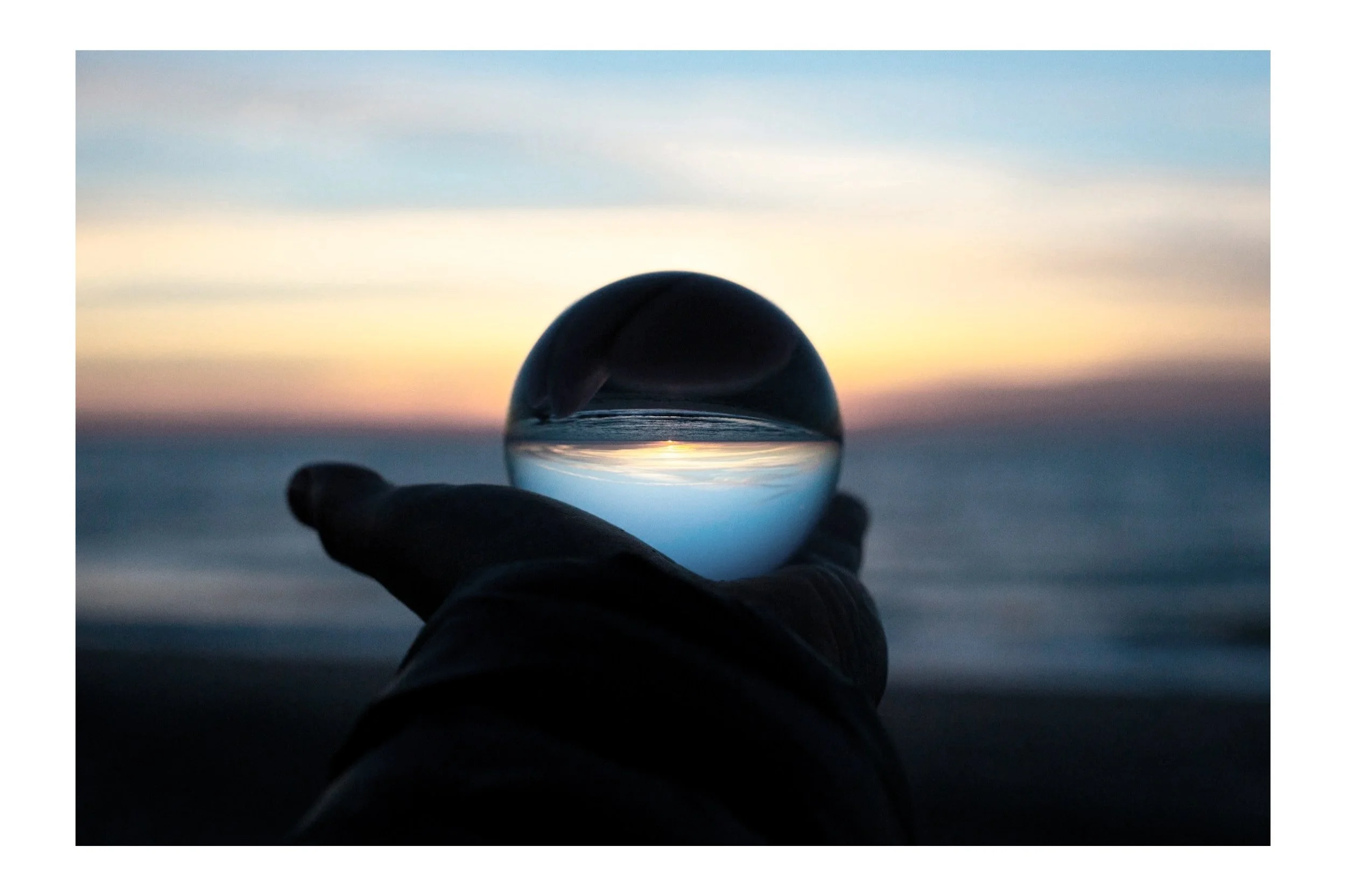 A hand holding a glass sphere on the beach during sunset, with the horizon and sky reflected upside down inside the sphere.