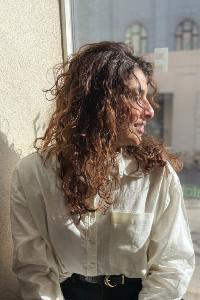 A woman with curly hair, wearing a cream-colored button-up shirt, is looking to her right at a sunlit window.