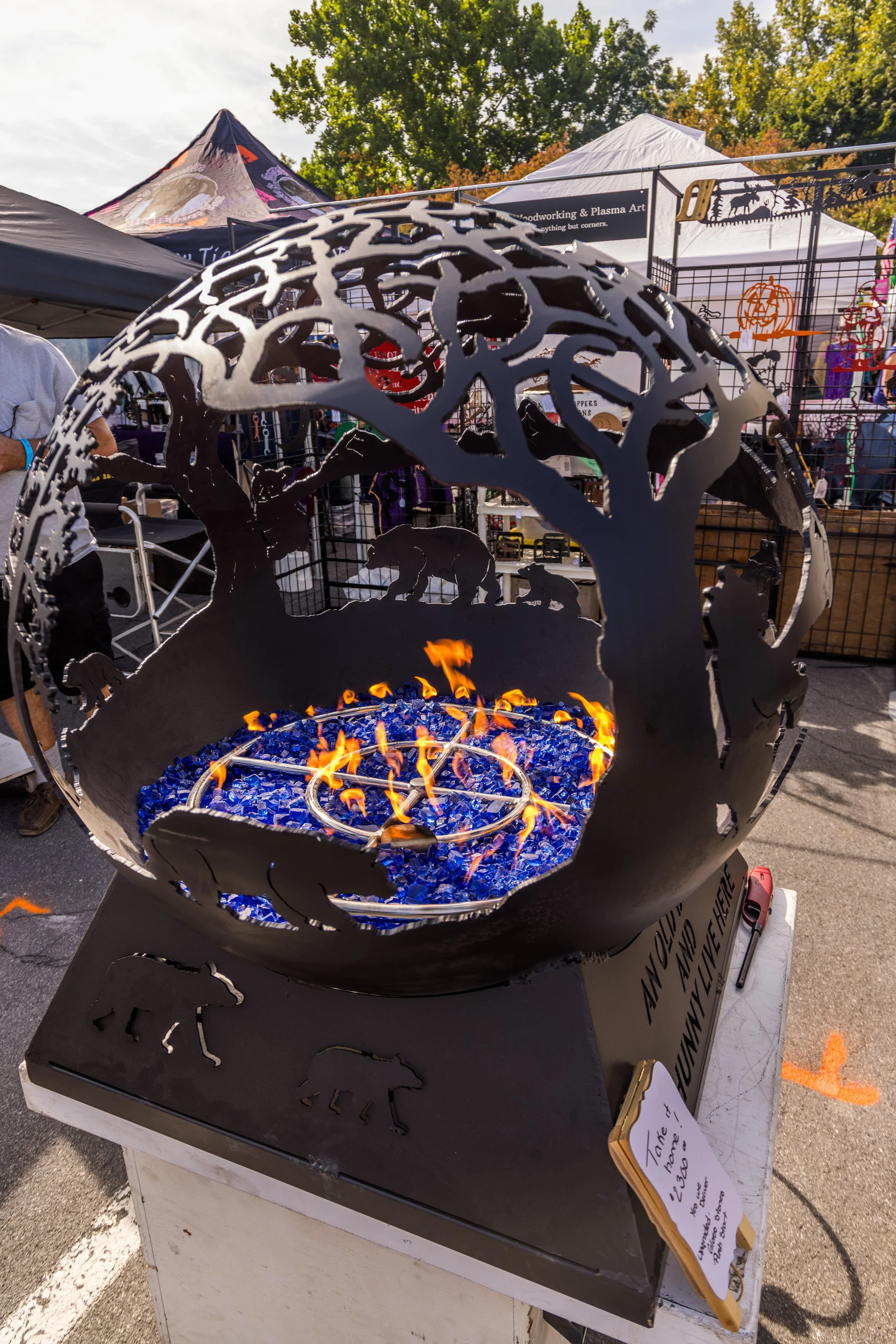 Decorative metal fire pit with animal and tree silhouette cutouts, burning blue electric rocks inside, displayed at an outdoor market.