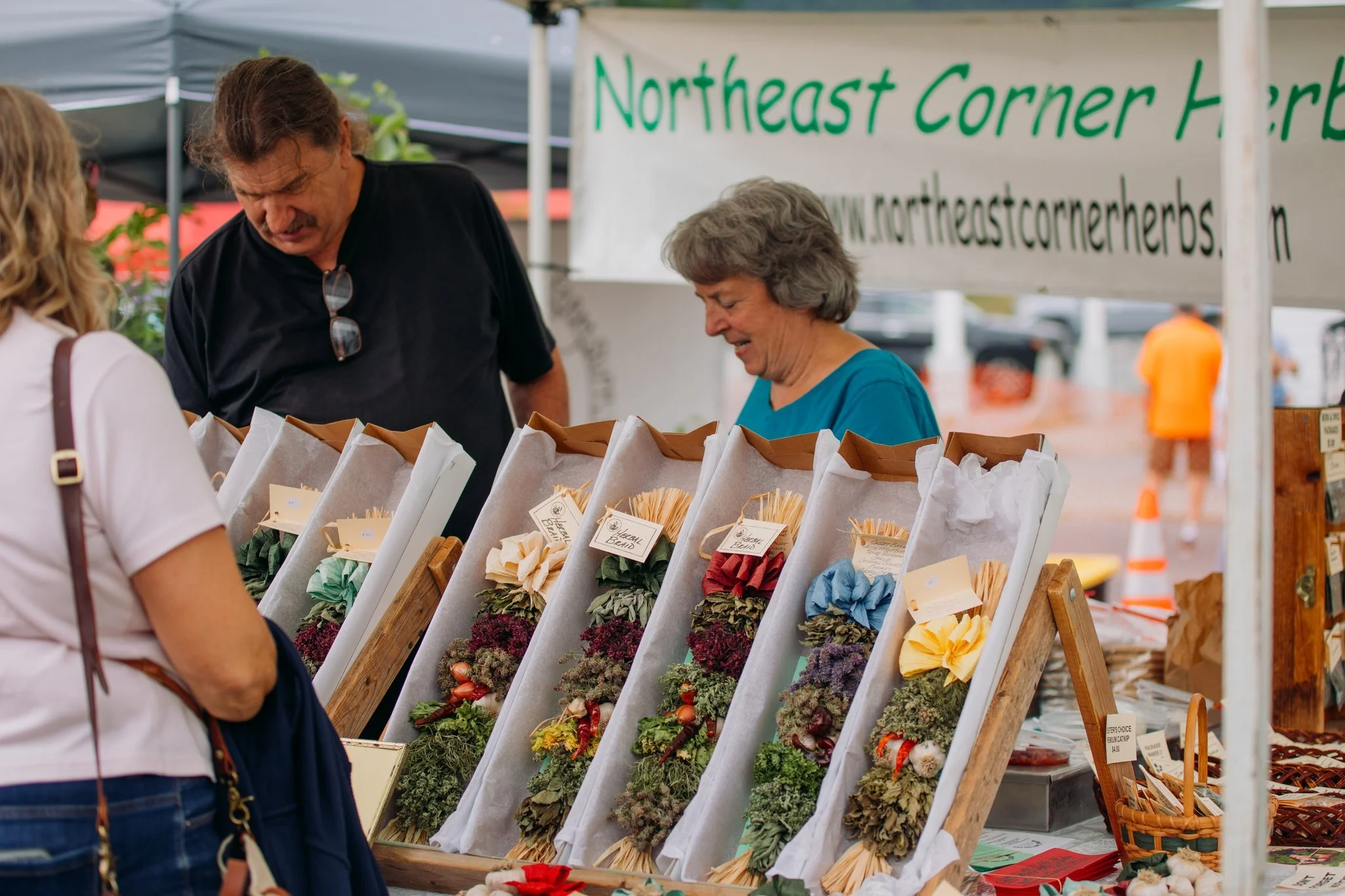 People shopping at an outdoor herb market booth with display of floral arrangements and herbs at Ottawa Northeast Corner Herbals market.