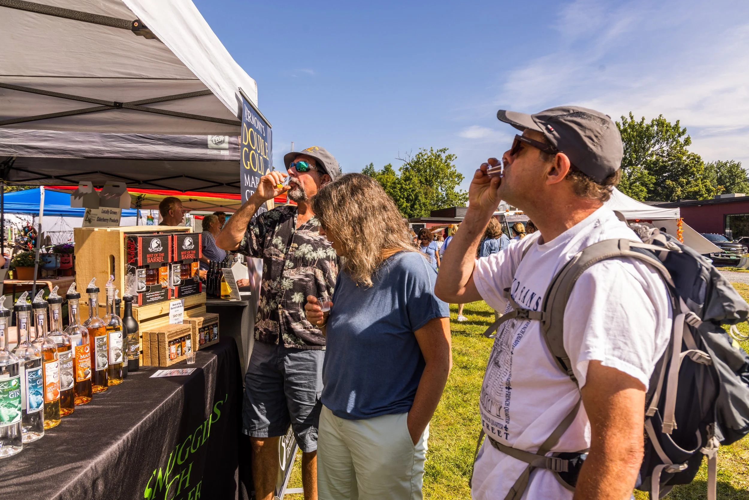 People at an outdoor market tasting whiskey at a booth with bottles on display, under a tent on a sunny day.