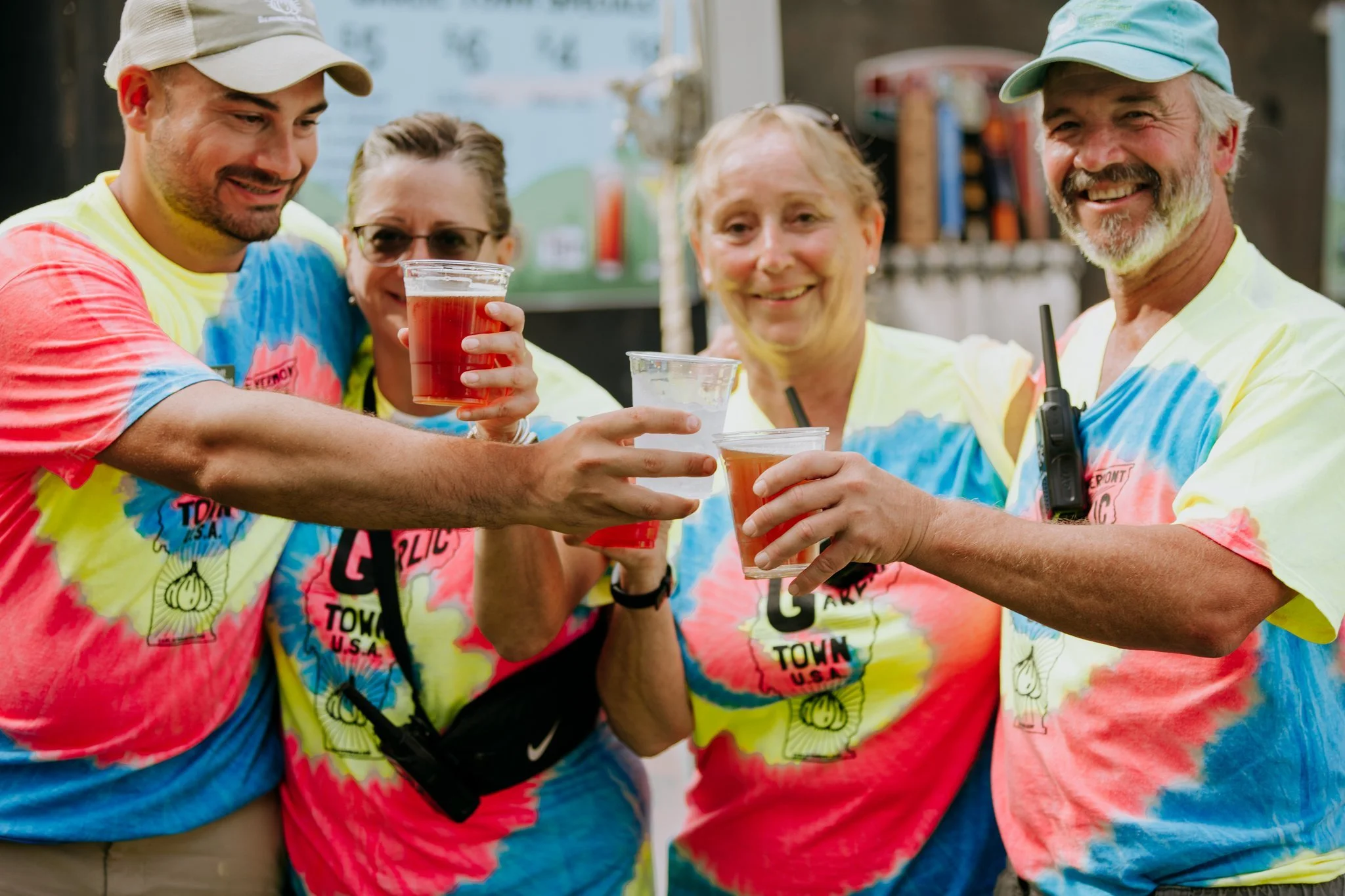 Group of four adults wearing tie-dye t-shirts, smiling, and raising plastic cups for a toast at an outdoor event.
