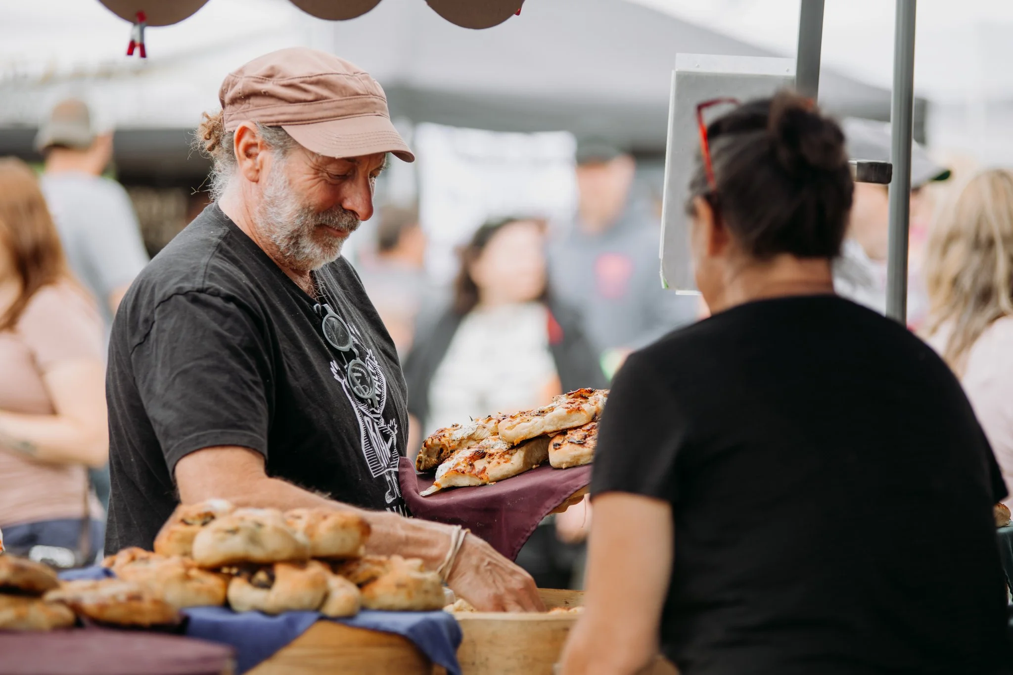 A man with a beard and glasses hanging around his neck is selling pizza at an outdoor market, handing a slice to a customer.