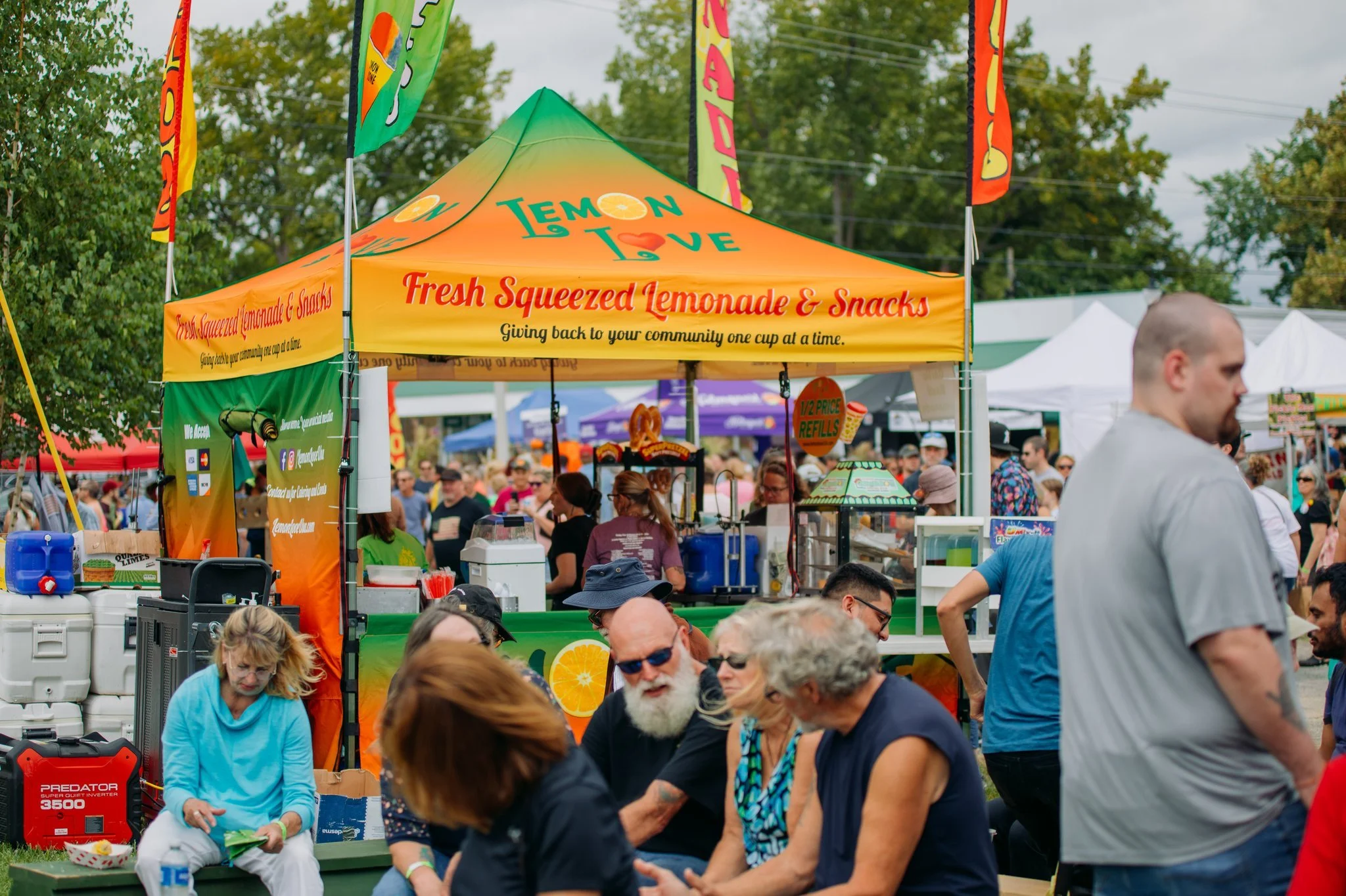 A crowded outdoor festival with a bright yellow and orange lemonade stand tent selling fresh squeezed lemonade and snacks, surrounded by people, trees, and other vendor tents.