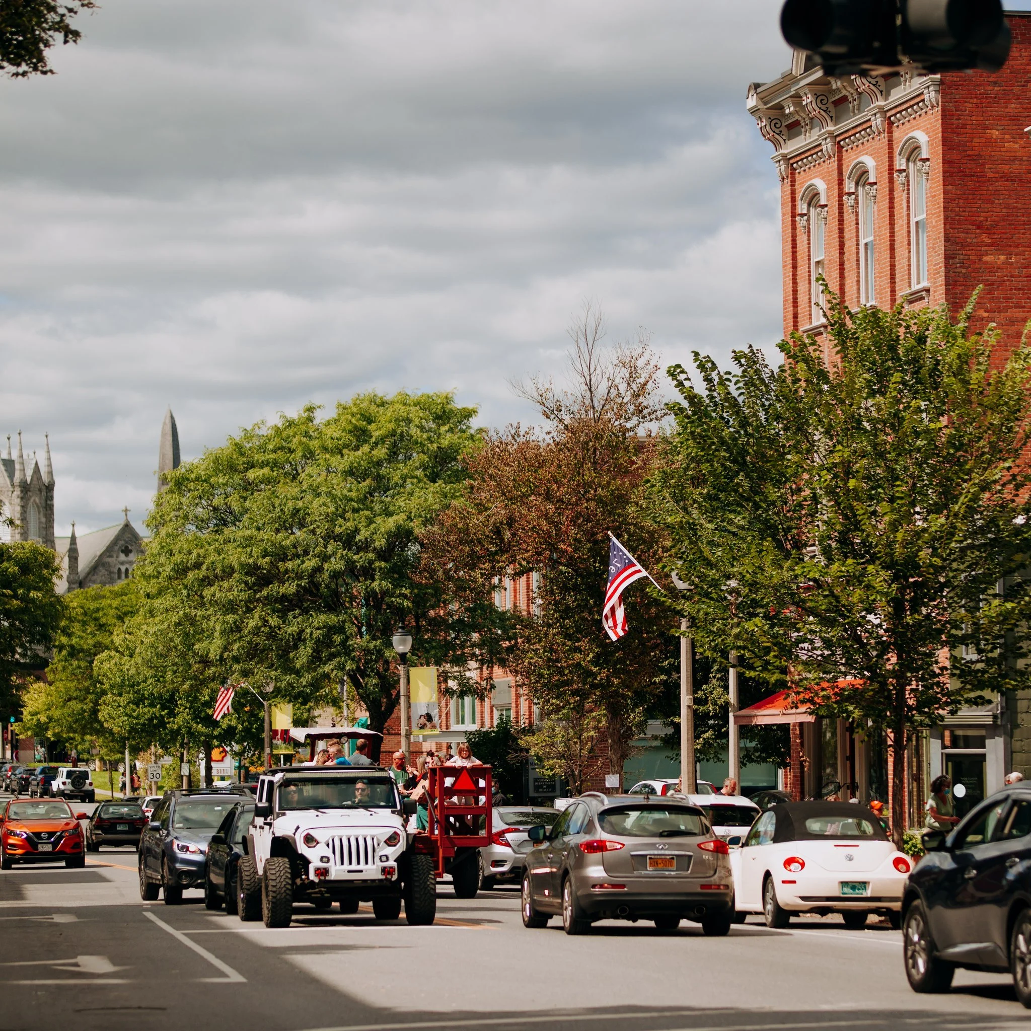 A busy street scene in a small town, with cars parked along the curb and a white Jeep driving through. People are walking on the sidewalk, some near storefronts and trees with green and brown leaves. An American flag is visible hanging from a building, and there are historic brick buildings and a church steeple in the background under a cloudy sky.
