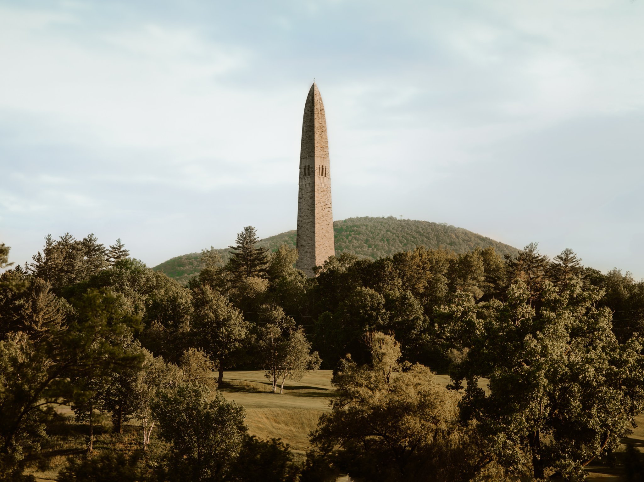 The Washington Monument standing tall amid lush green trees and hills under a partly cloudy sky.