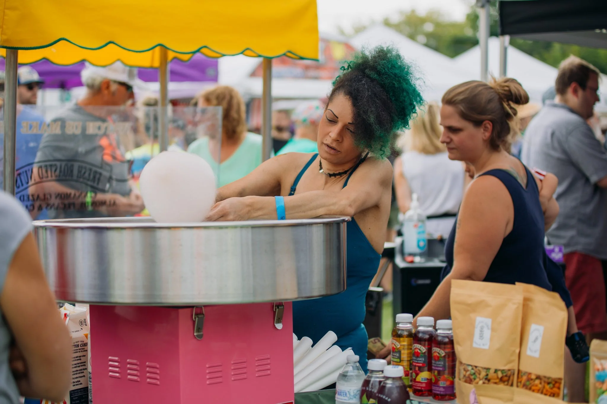 A woman with curly green hair making cotton candy at an outdoor fair or market, surrounded by other people and tents.