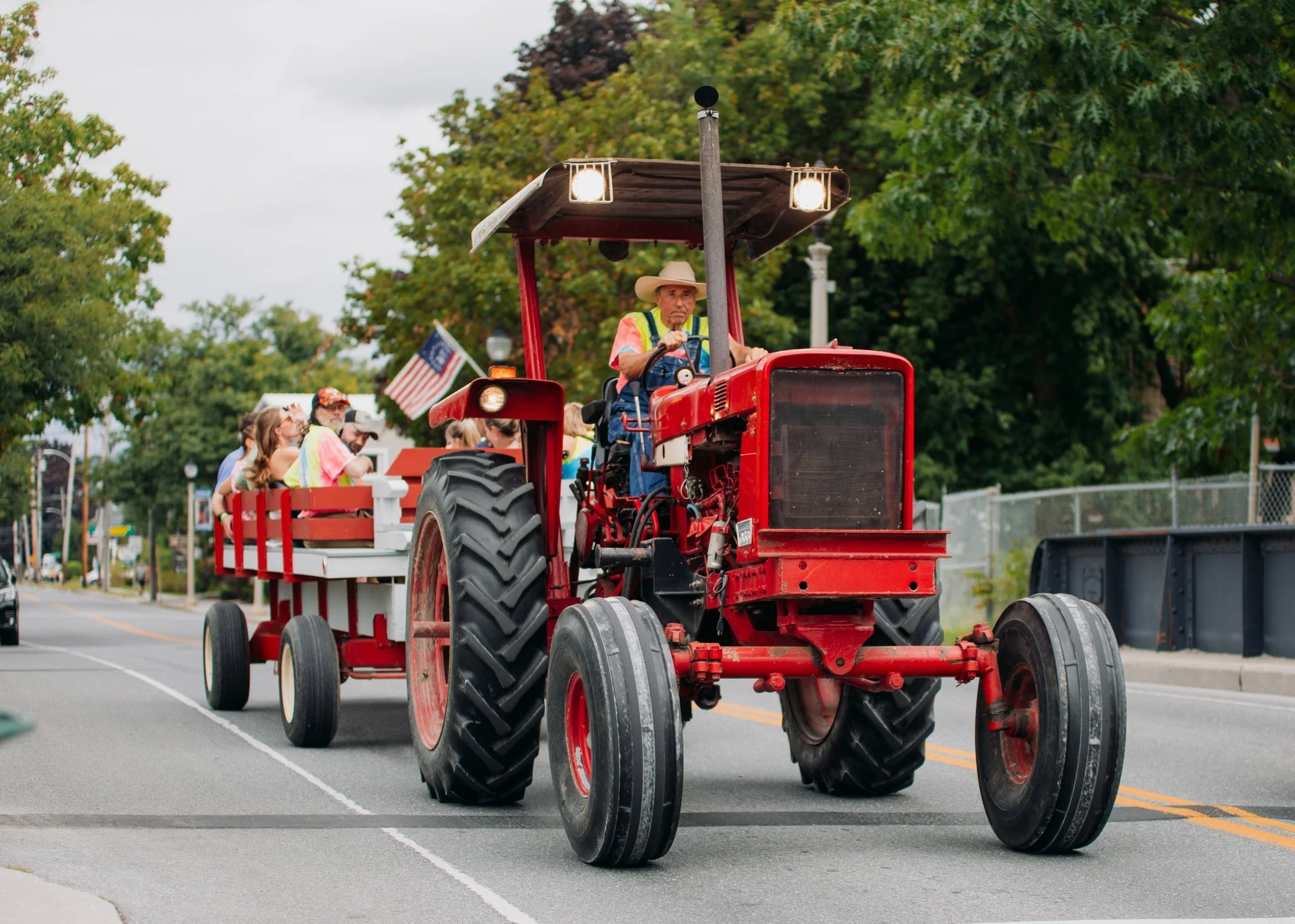 A man in a cowboy hat drives a red tractor pulling a float with people sitting in it, during a parade on a street lined with trees and American flags.