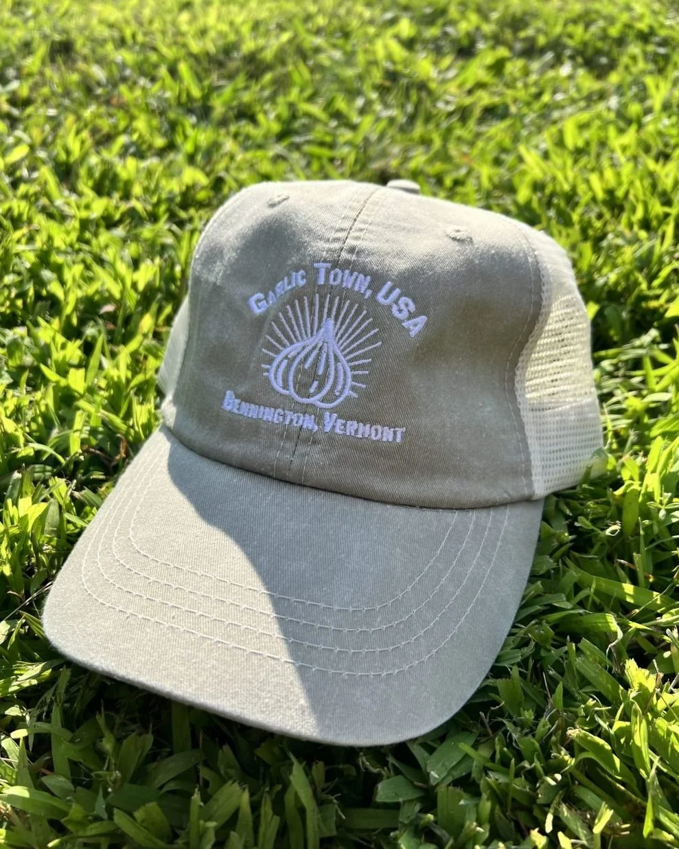 A beige trucker cap with white embroidery that reads 'Garlic Town, USA' and 'Brownington Vermont', with a garlic bulb illustration, resting on green grass.