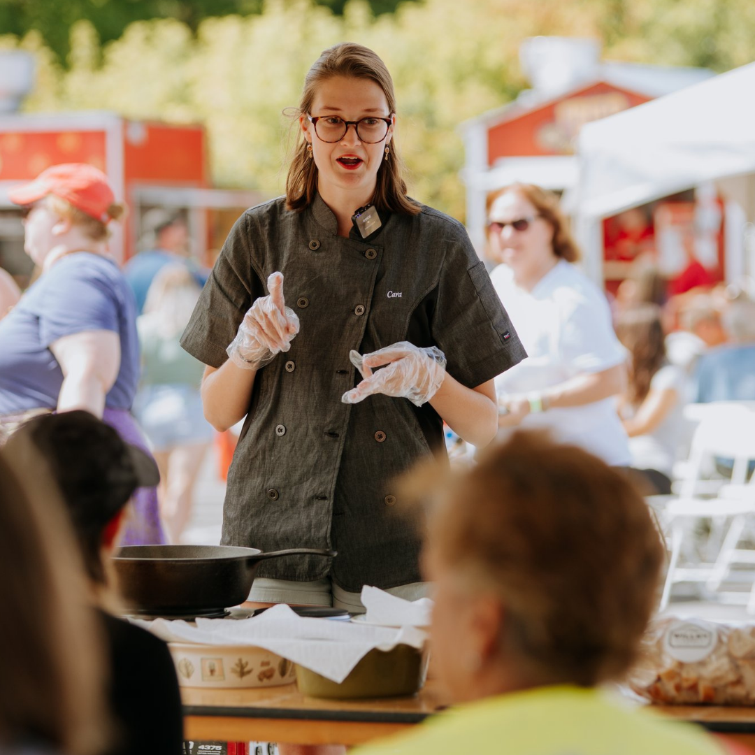 A woman with glasses, wearing a chef's coat and gloves, giving a cooking demonstration at an outdoor event with people watching.