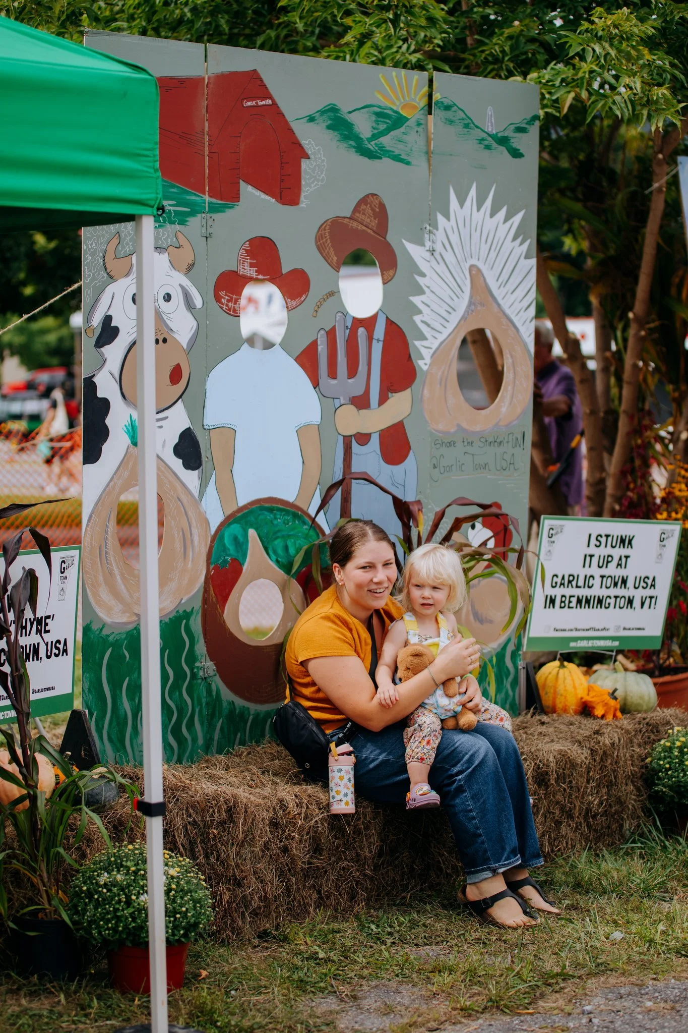 A woman holding a young girl sitting on a hay bale in front of a decorated booth at a fall festival, with a painted scene of a cow and people, pumpkins, and fall decorations nearby.