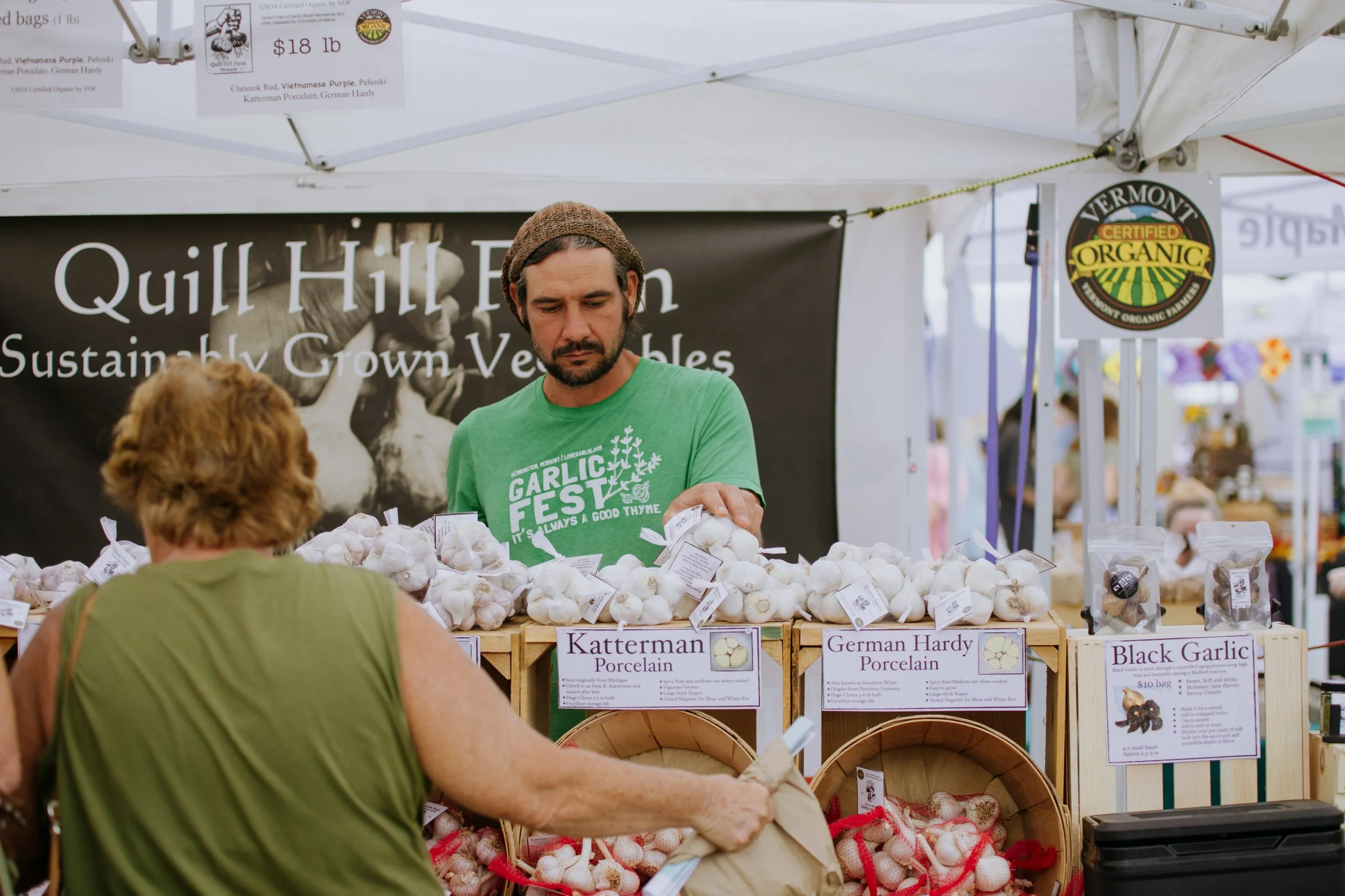 A man with a beard wearing a green T-shirt and a brown beanie sells garlic bulbs at an outdoor farmers market booth. A woman with short, curly hair and a sleeveless green top shops at the booth. The booth displays labels for garlic varieties, includi