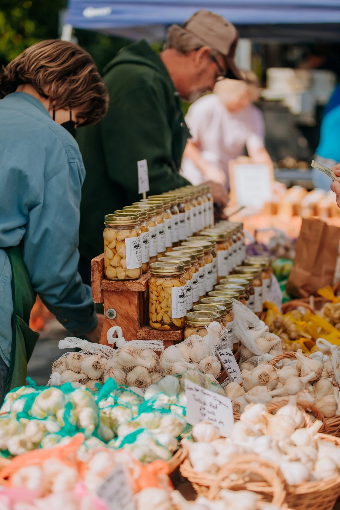People shopping at an outdoor farmers market with jars of preserved garlic and bulbs of garlic on display.