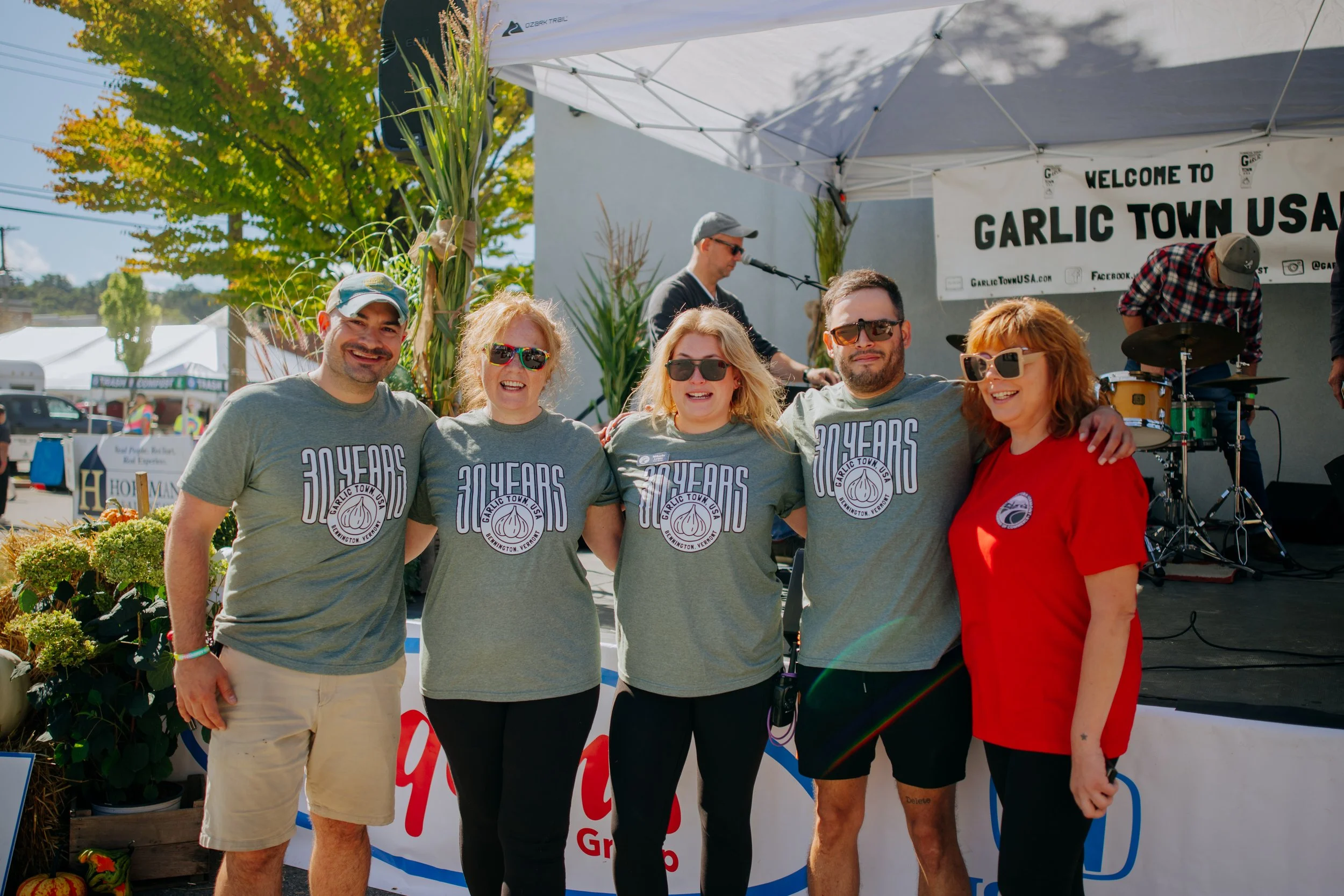 Group of five people standing together in front of a stage at a festival, with a band playing in the background under a banner that says 'Welcome to Garlic Town USA'.