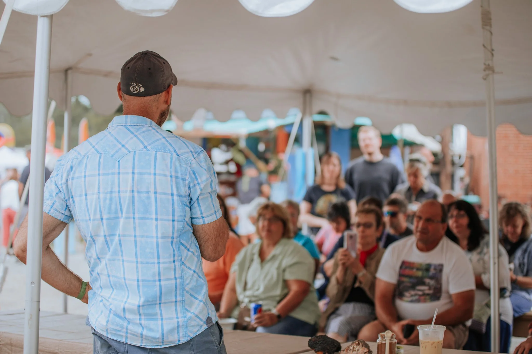 A man with a baseball cap and light blue plaid shirt standing at a booth speaking to a seated group of people under a tent at an outdoor event.
