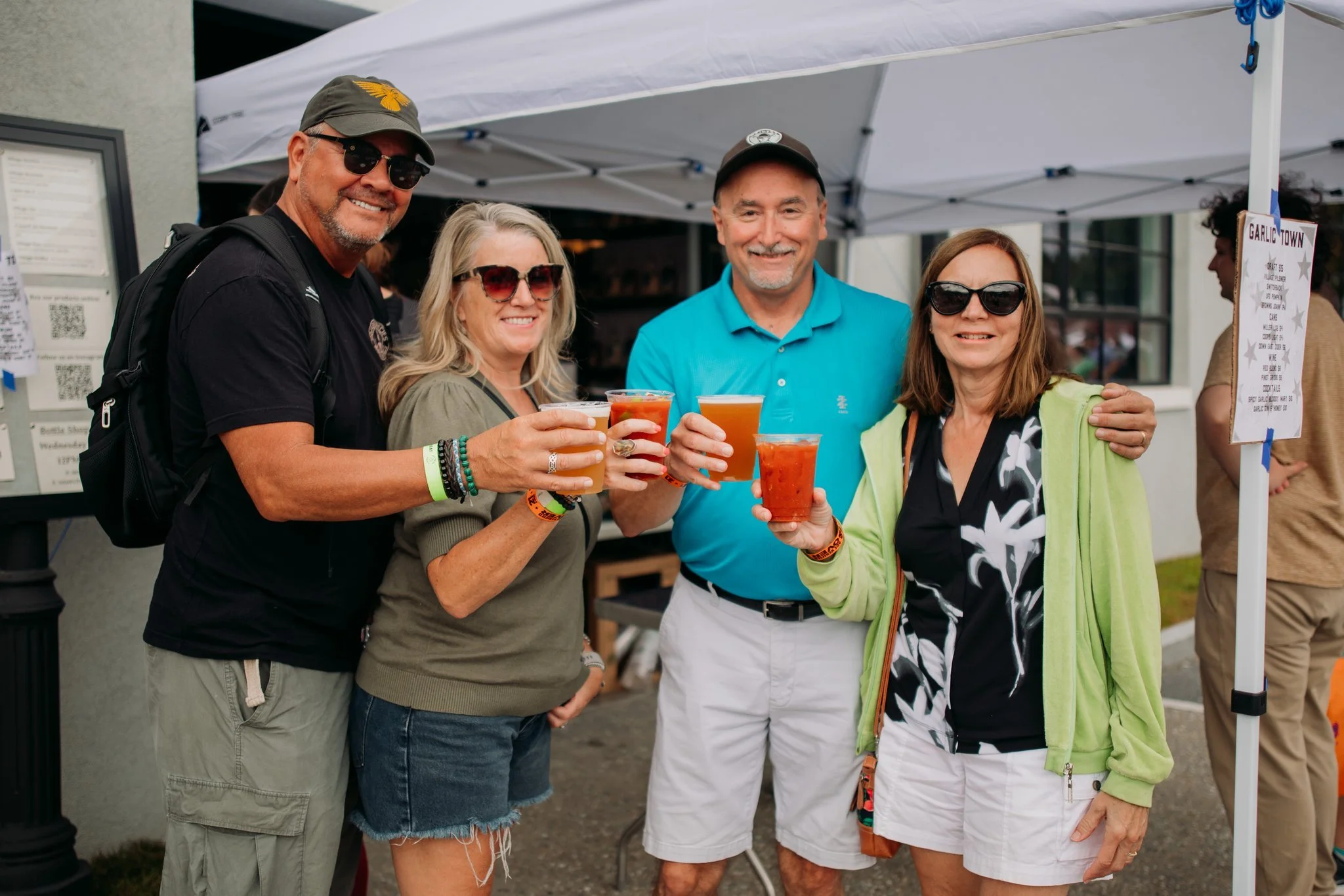 Four people are standing outdoors under a white canopy, holding drinks and smiling. They are dressed casually with sunglasses, and there is a sign to the right that reads 'Garlic Town'.