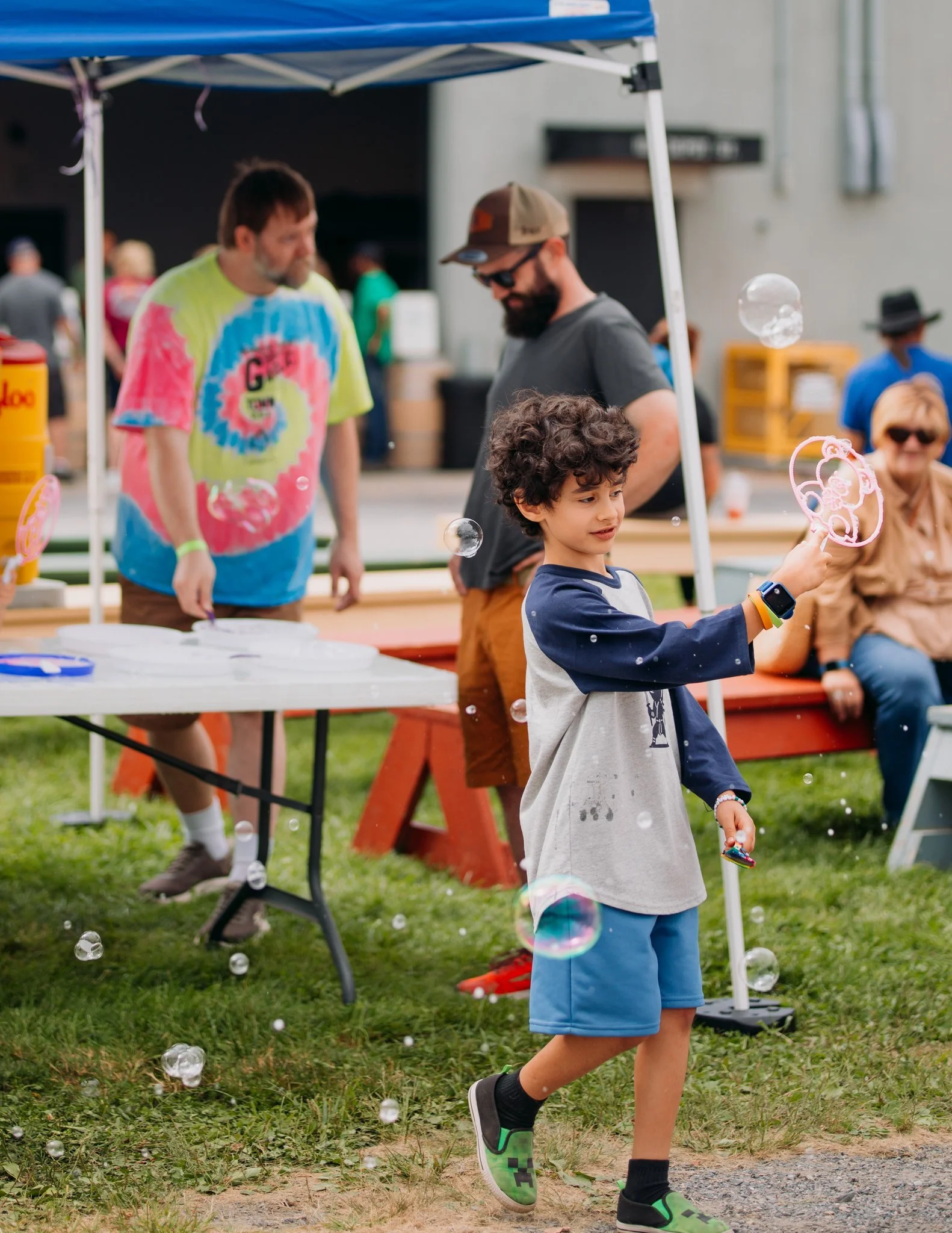 A young boy with curly hair playing with bubbles at an outdoor event, with people visible in the background under a blue canopy and sitting on a bench.