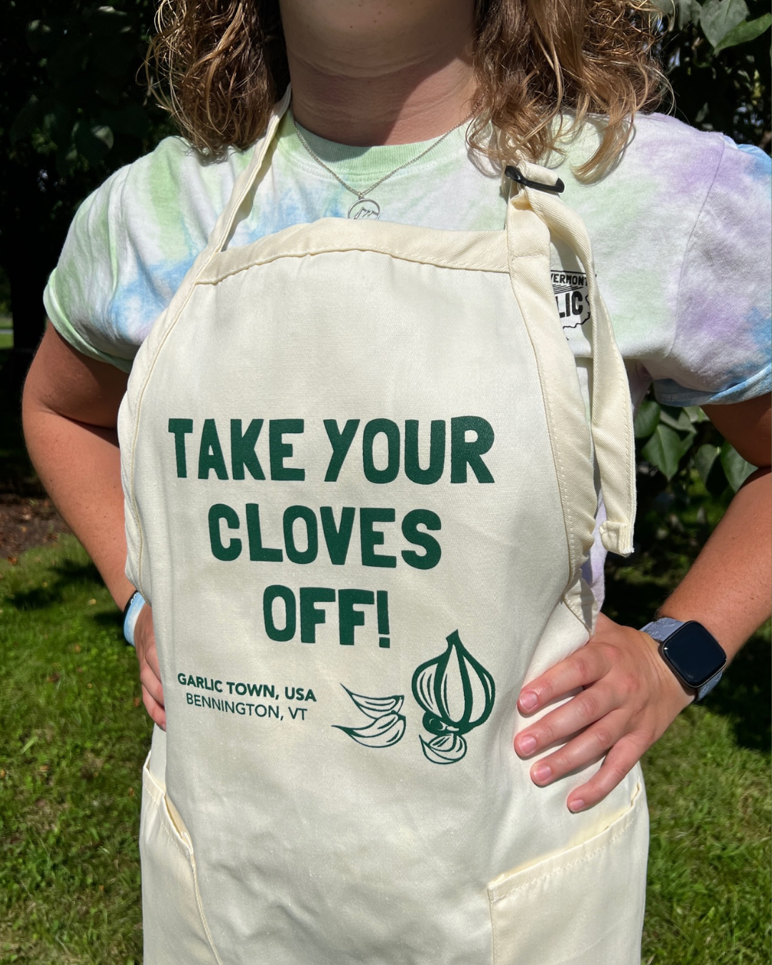 A person wearing a tie-dye t-shirt, an apron with the text "Take Your Gloves Off!" and location details, standing outdoors with green grass and bushes in the background.