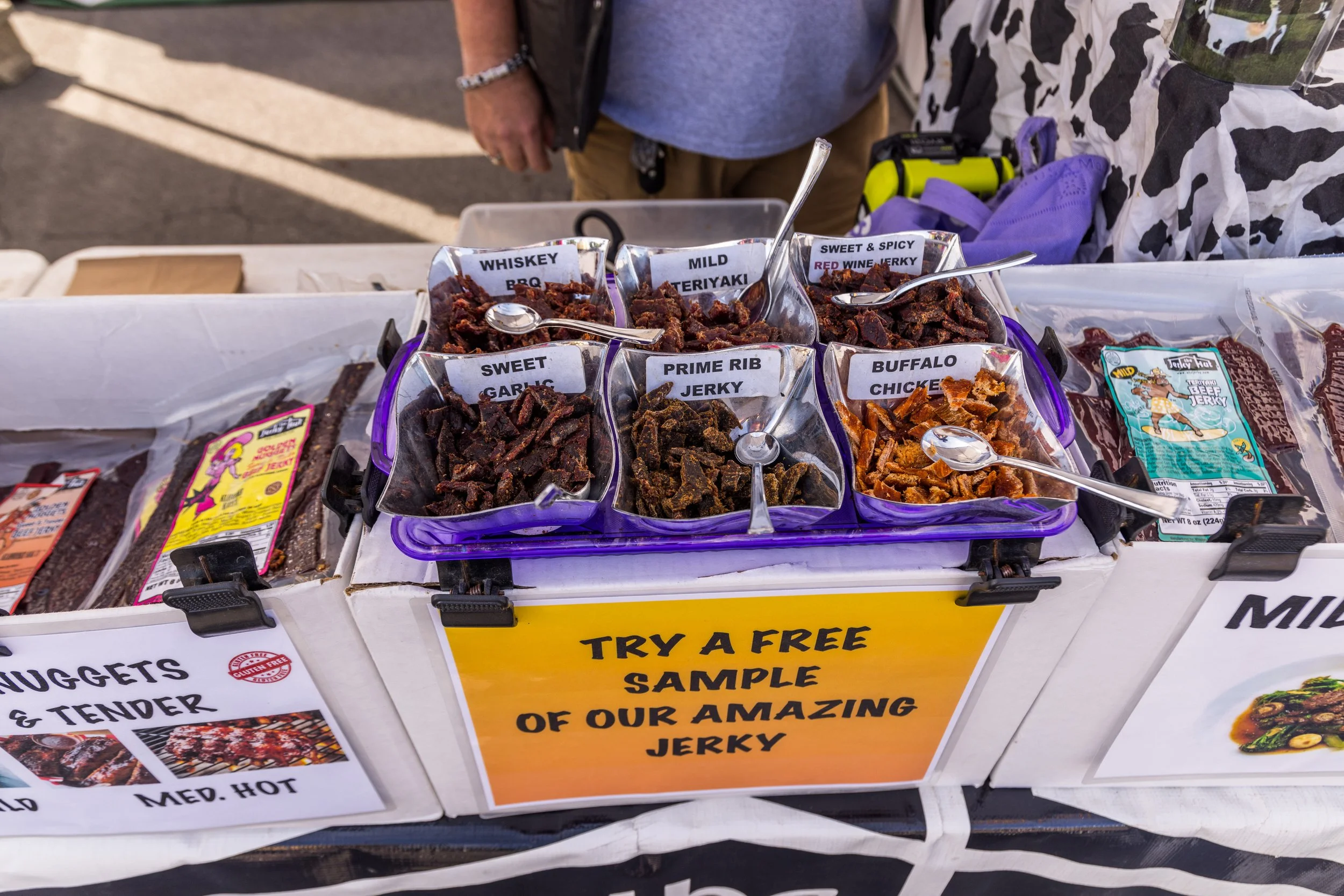 A table displaying various types of beef jerky in small containers with labeled signs, including whiskey BBQ, mild teriyaki, sweet & spicy red wine jerky, sweet garlic, prime rib jerky, and buffalo chicken. A yellow sign promotes free sampling.