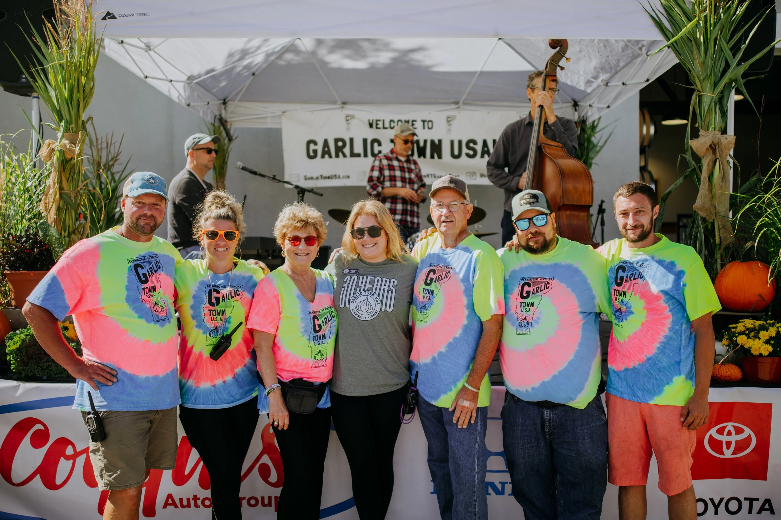 A group of seven people wearing colorful tie-dye shirts with their arms around each other, standing in front of a stage with musicians playing instruments. The stage has a banner reading 'Welcome to Garlic Town USA,' and there are plants and pumpkins