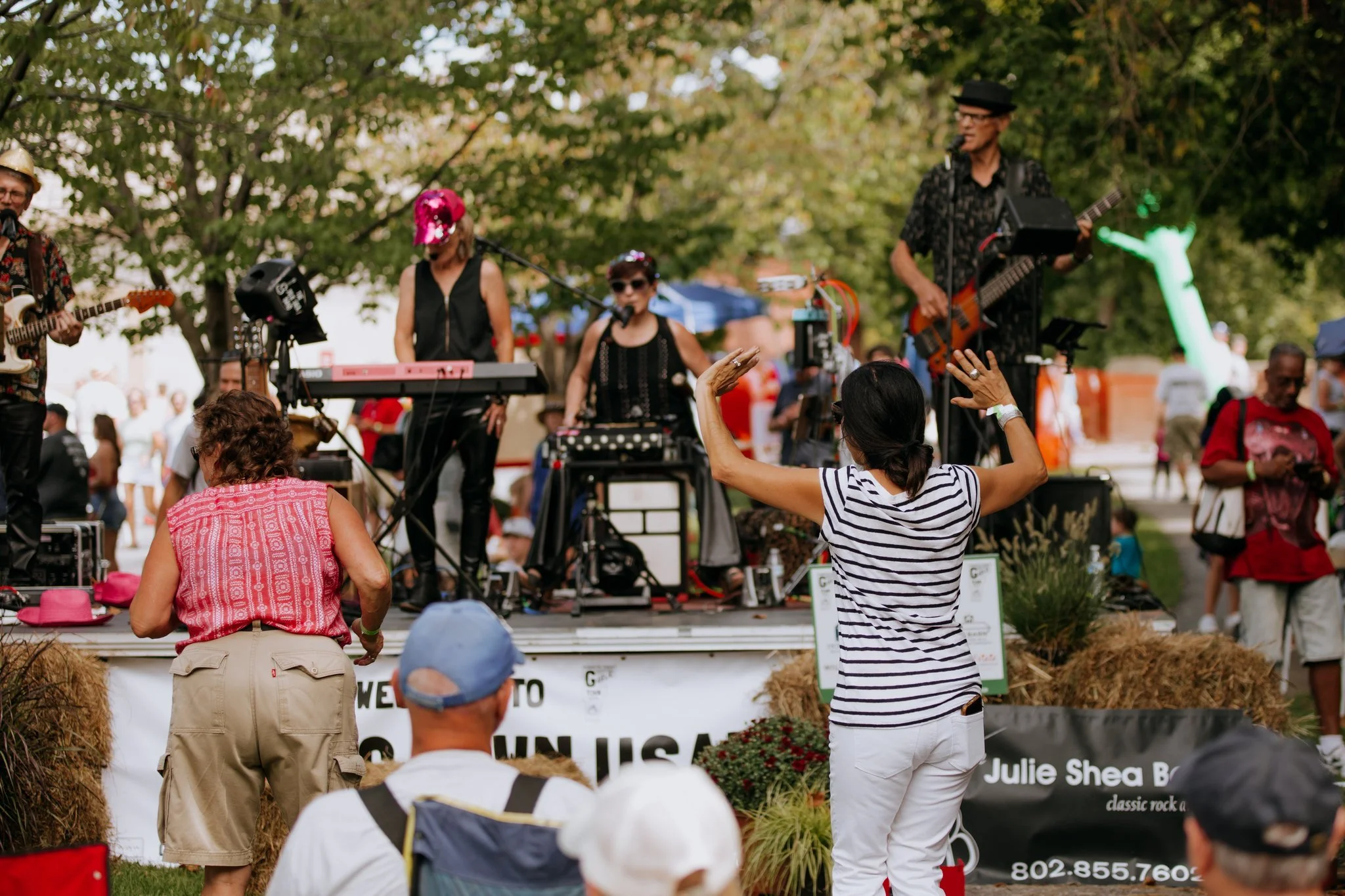 A woman in a striped shirt dancing in front of a live band playing outdoors. The band members are on a stage with various instruments, and there are other people watching and enjoying the music.