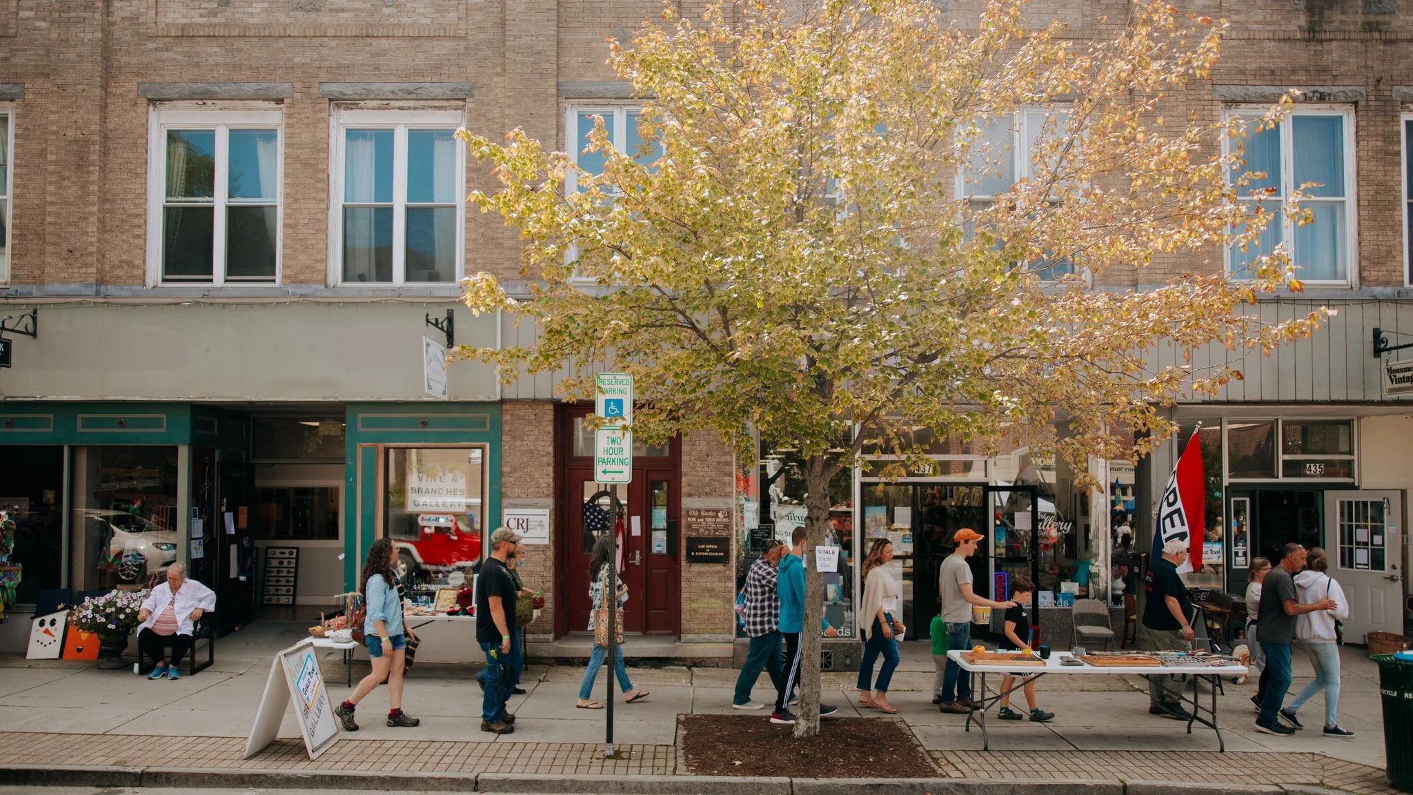 People walking on sidewalk in a small town with storefronts, a large tree with fall foliage, and two parking signs, one for reserved handicapped parking and one for two-hour parking.
