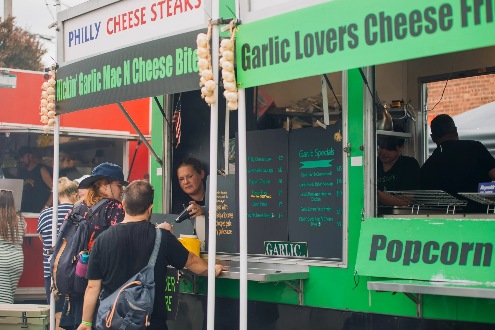 People ordering food at a green food truck with a menu board displaying garlic special items.