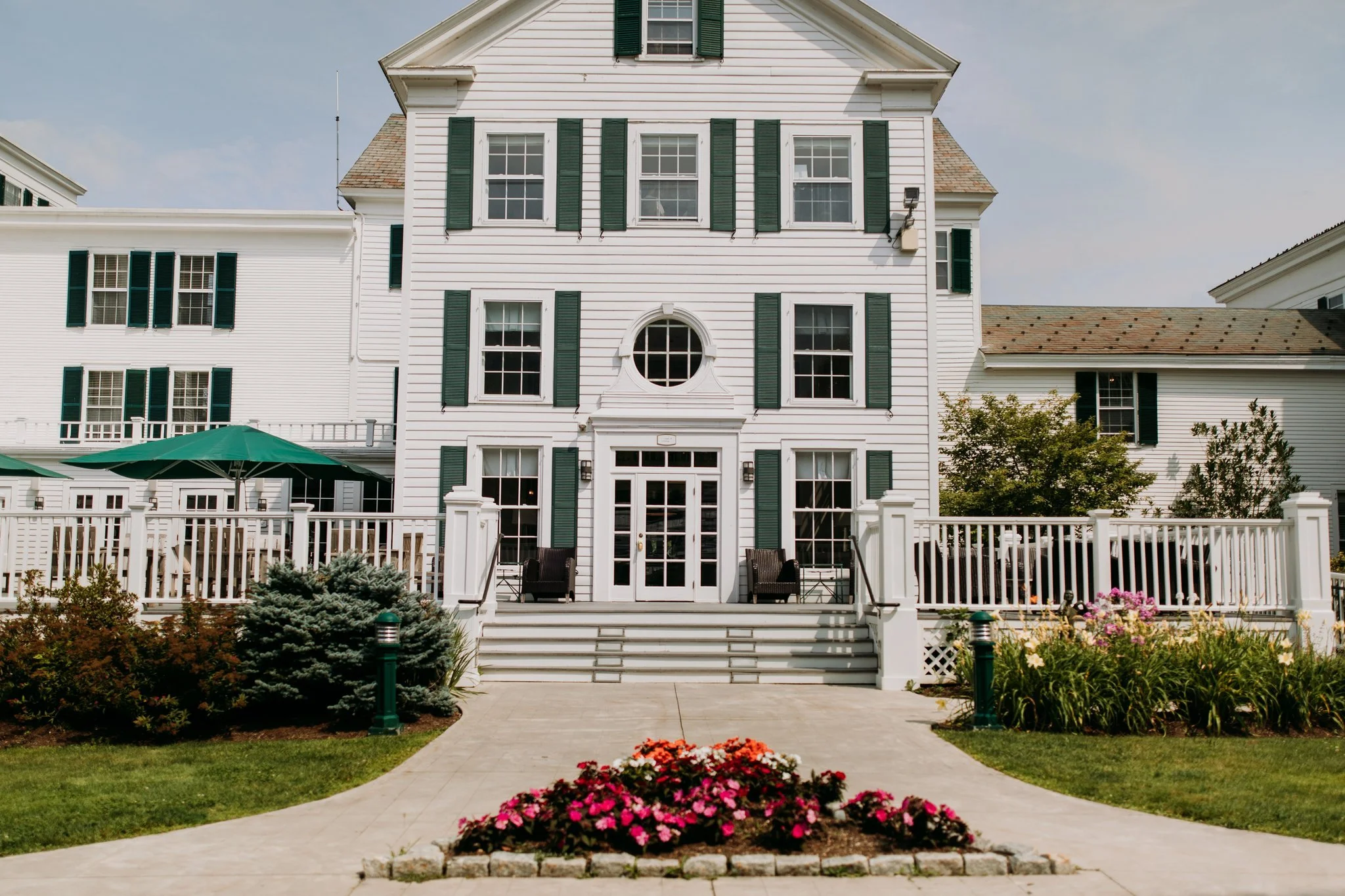 A white multi-story house with dark green shutters and a front porch with stairs, surrounded by a garden with flowers and bushes, under a partly cloudy sky.