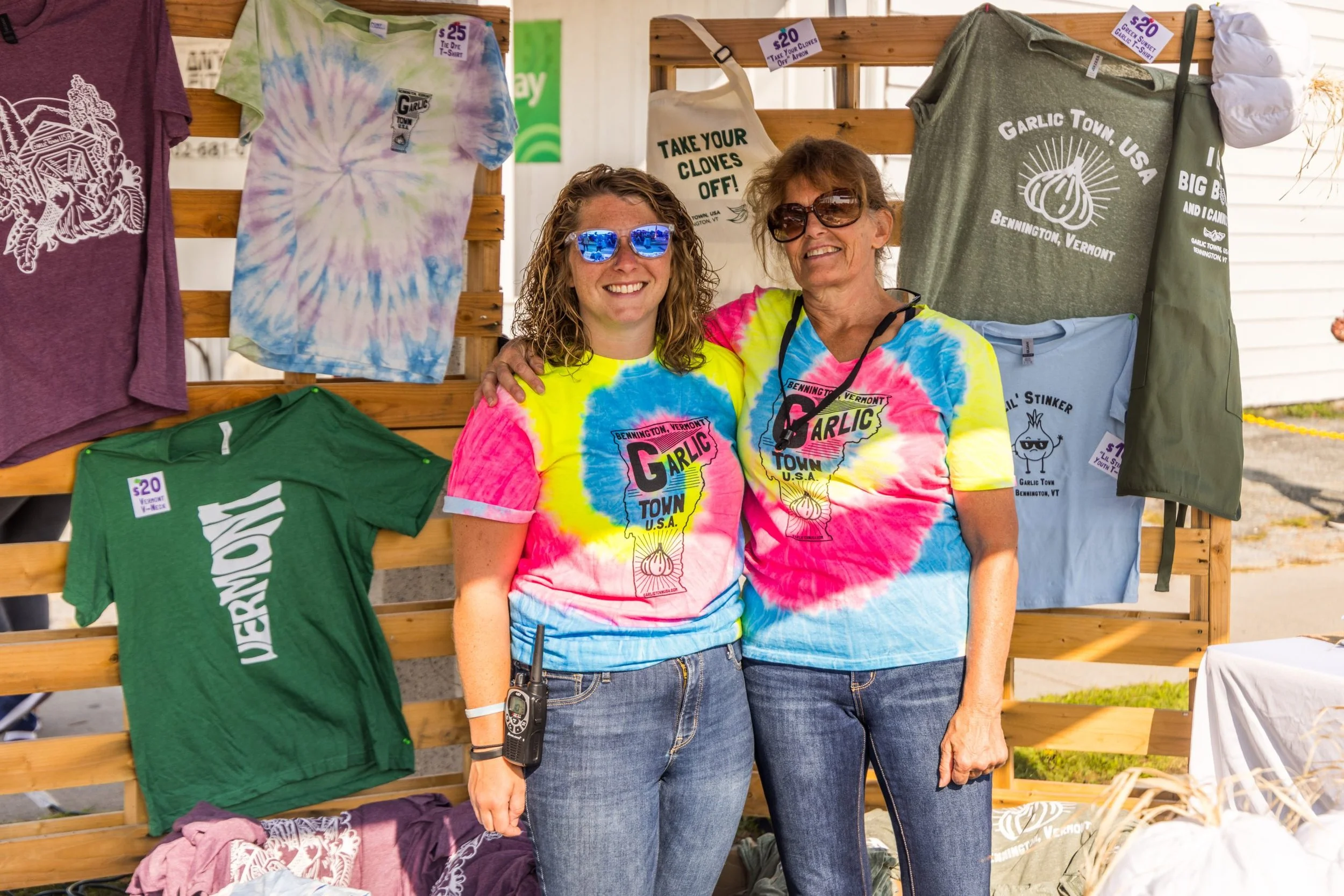 Two women smiling at a booth selling tie-dye T-shirts and other apparel at an outdoor event. They are wearing matching colorful tie-dye shirts with the event's logo, and there are various shirts hanging on a wooden display behind them.