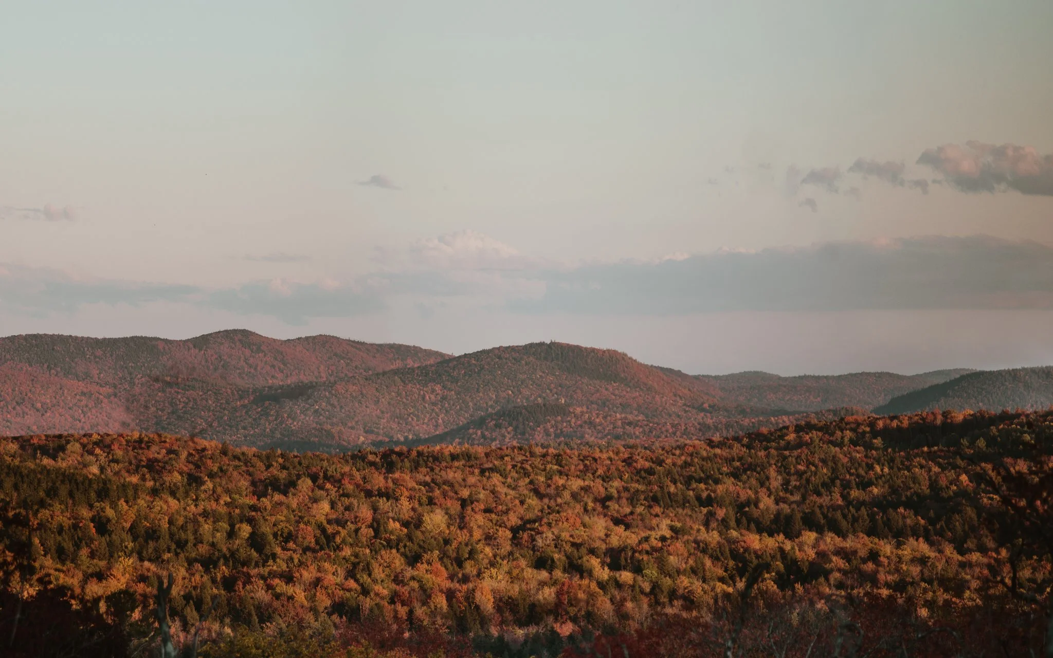 Scenic landscape of rolling hills and mountains covered with autumn trees under a mostly cloudy sky.