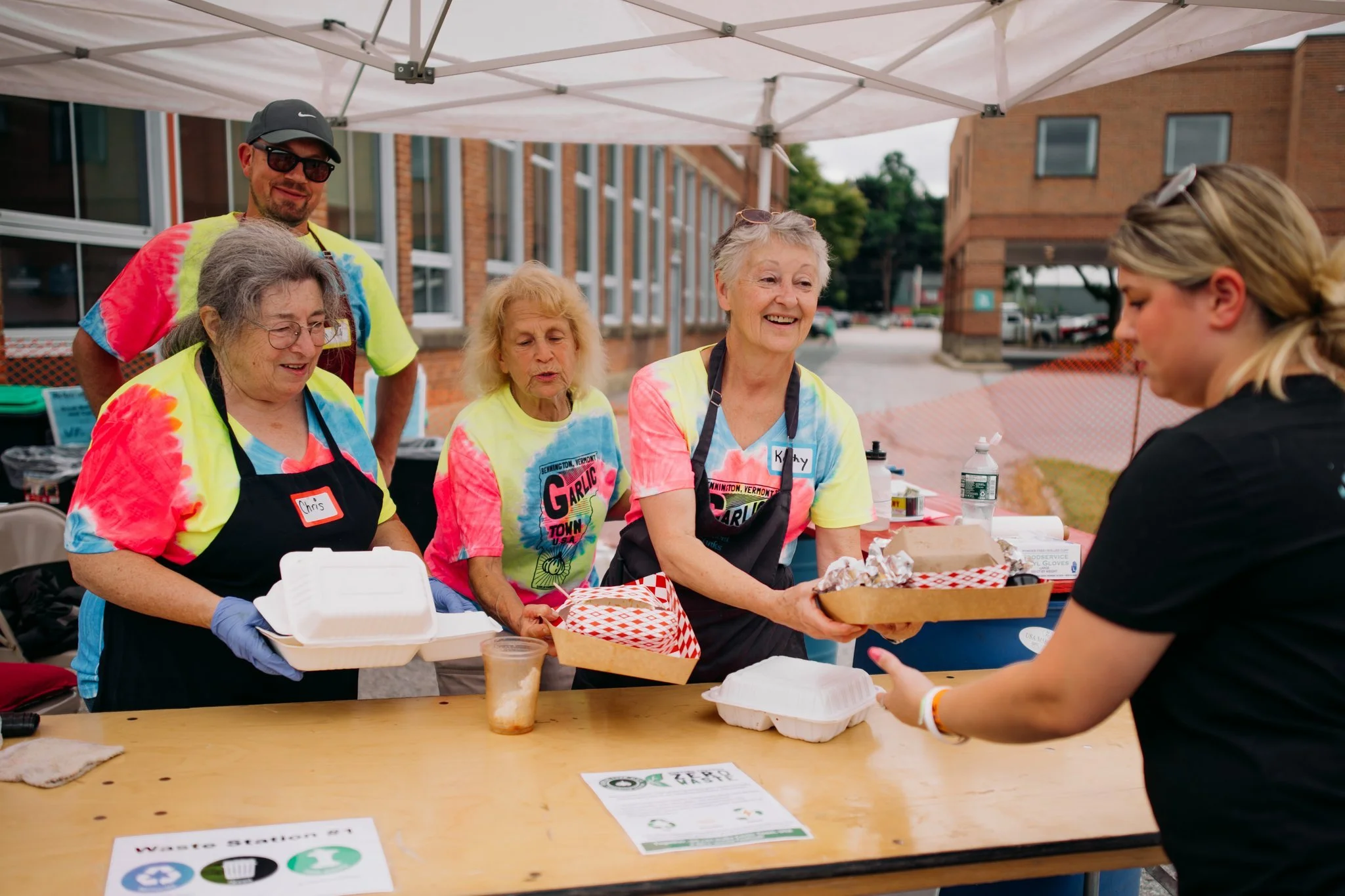 Group of five adults volunteer at a food distribution stand under a canopy, handing food containers to a woman. They are wearing colorful tie-dye shirts.