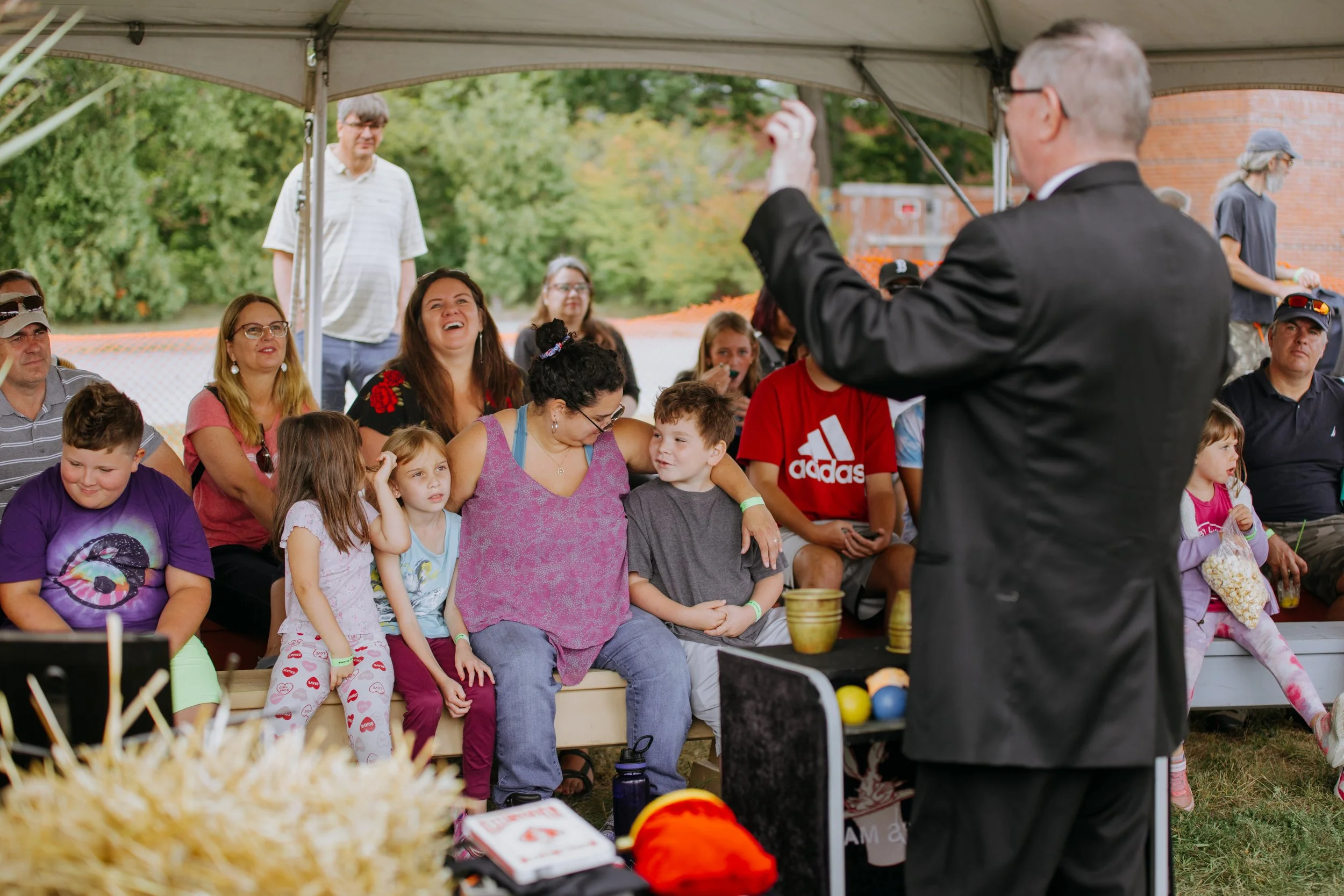 A man in a suit performing a magic trick for a group of children and adults under a tent at an outdoor event.