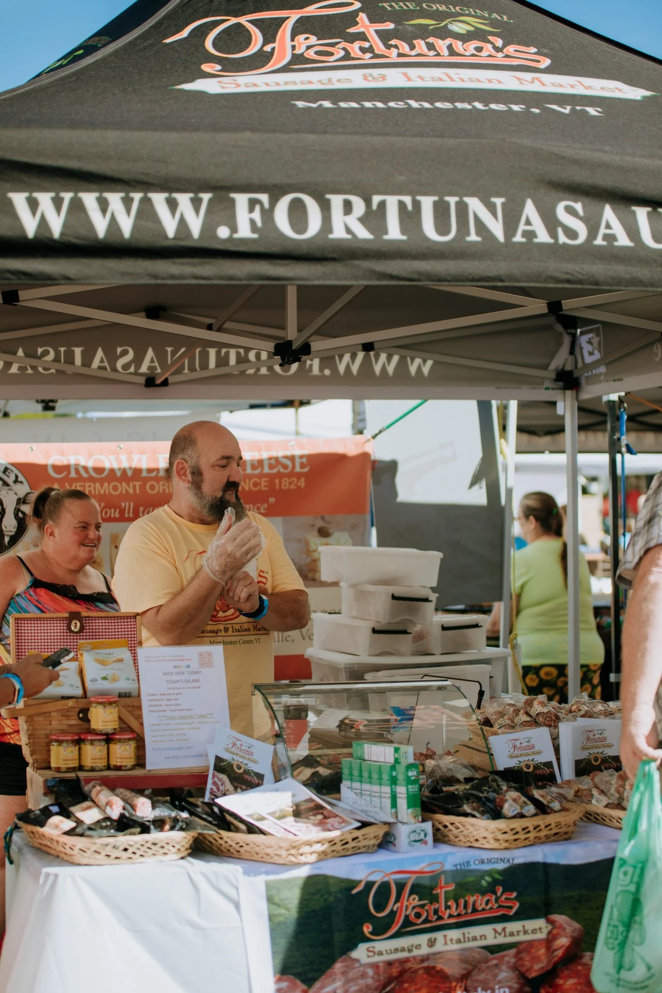 People at a market stall selling sausage and Italian meats, with a vendor preparing food and customers browsing the products.