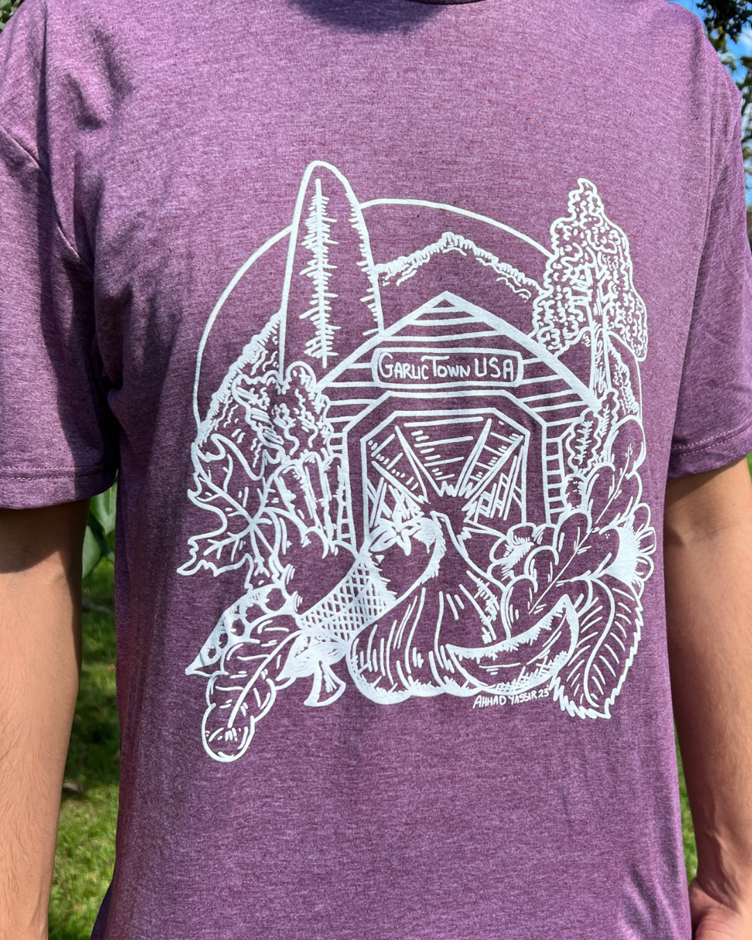 Purple T-shirt with a white line art design of a landscape featuring mountains, trees, a waterfall, and a wooden arch with the words 'Garlic Town USA' on it.