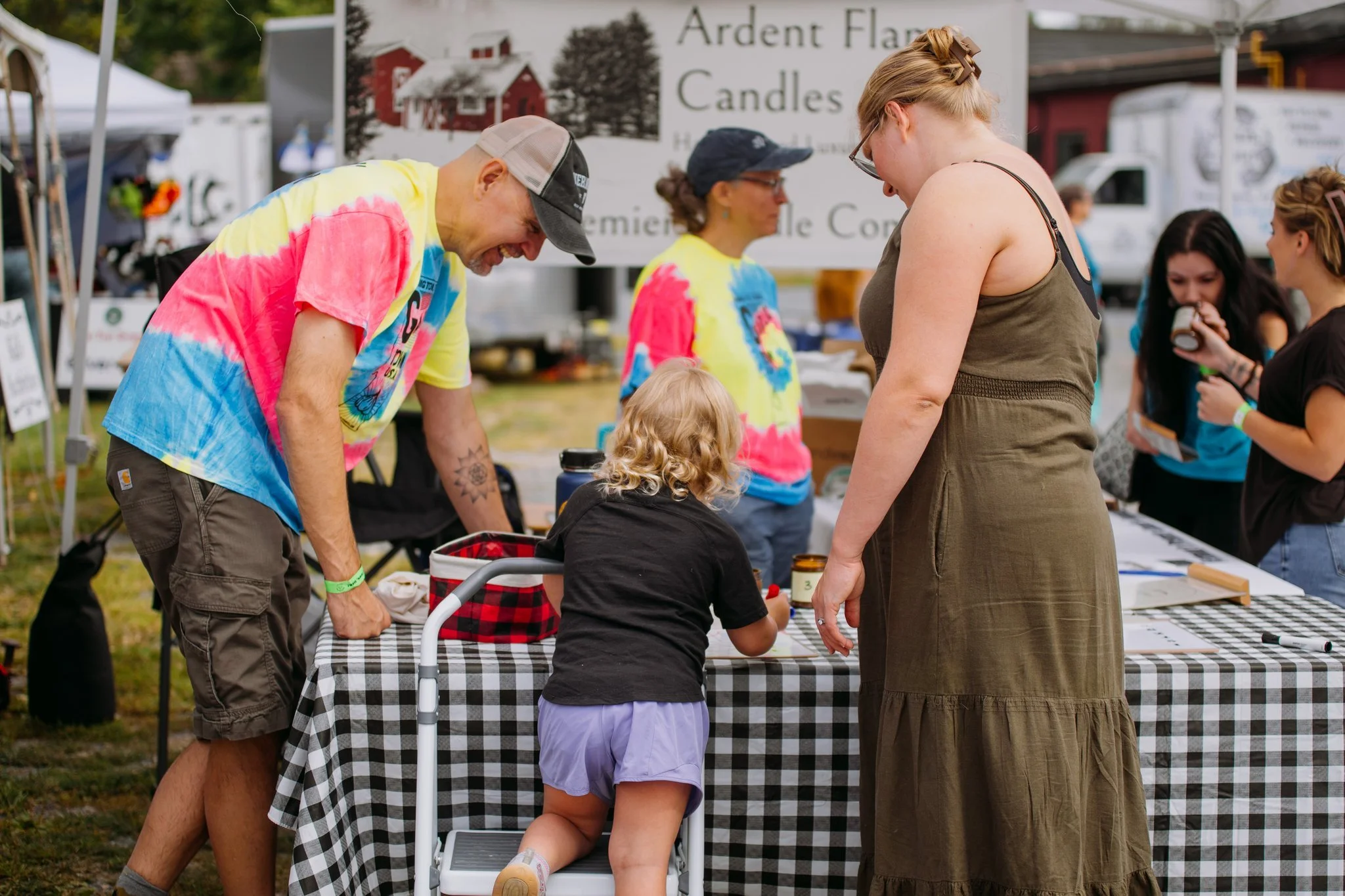 People gathered at an outdoor market booth with a black-and-white checkered tablecloth, talking and looking at items, with a child leaning on the table and a woman wearing glasses and a brown dress in front.