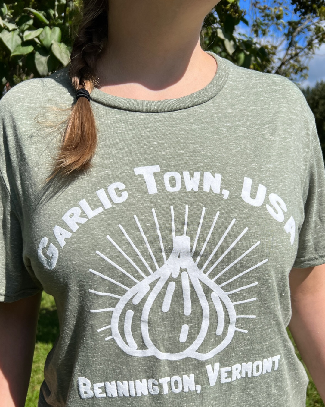 A person wearing a green T-shirt with white text and a graphic. The text reads 'Garlic Town, USA' and 'Bennington, Vermont.' The graphic features a stylized garlic bulb with radiating lines.