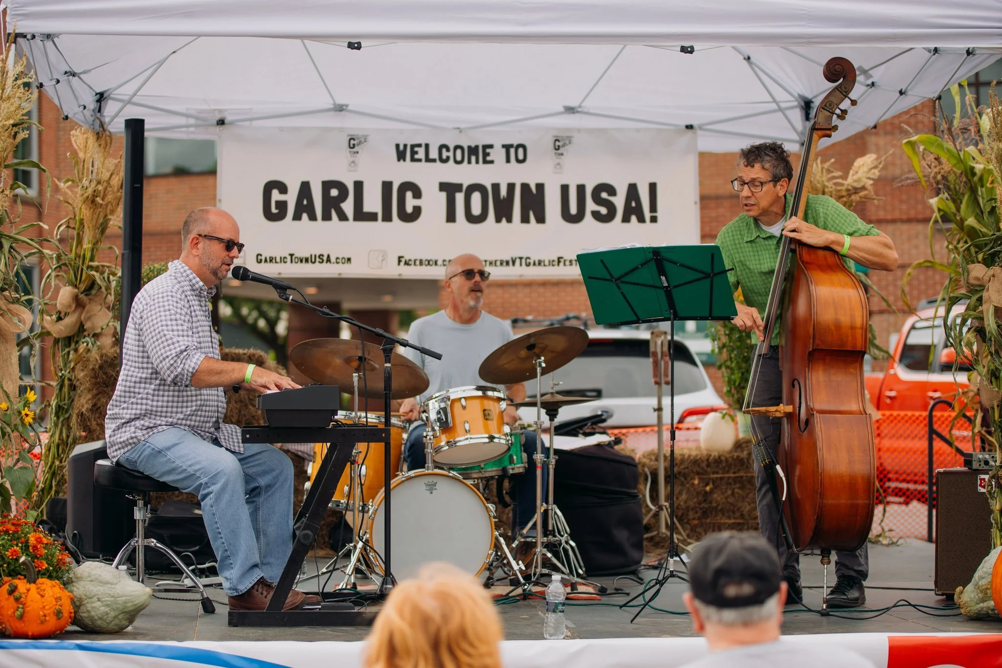 A three-piece band performing on stage at Garlic Town USA festival, featuring a keyboard player, drummer, and double bass player, with a sign reading 'Welcome to Garlic Town USA' in the background, and autumn decorations around the stage.