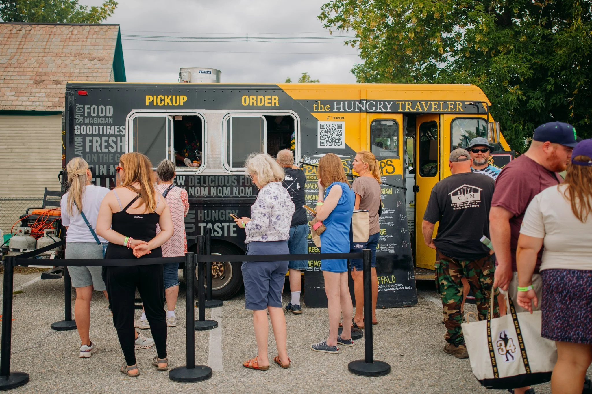 People standing in line at a food truck called the Hungry Traveler, which has a yellow and black exterior. The truck offers food pickup and orders, with menu items listed on the side and a QR code for scanning.