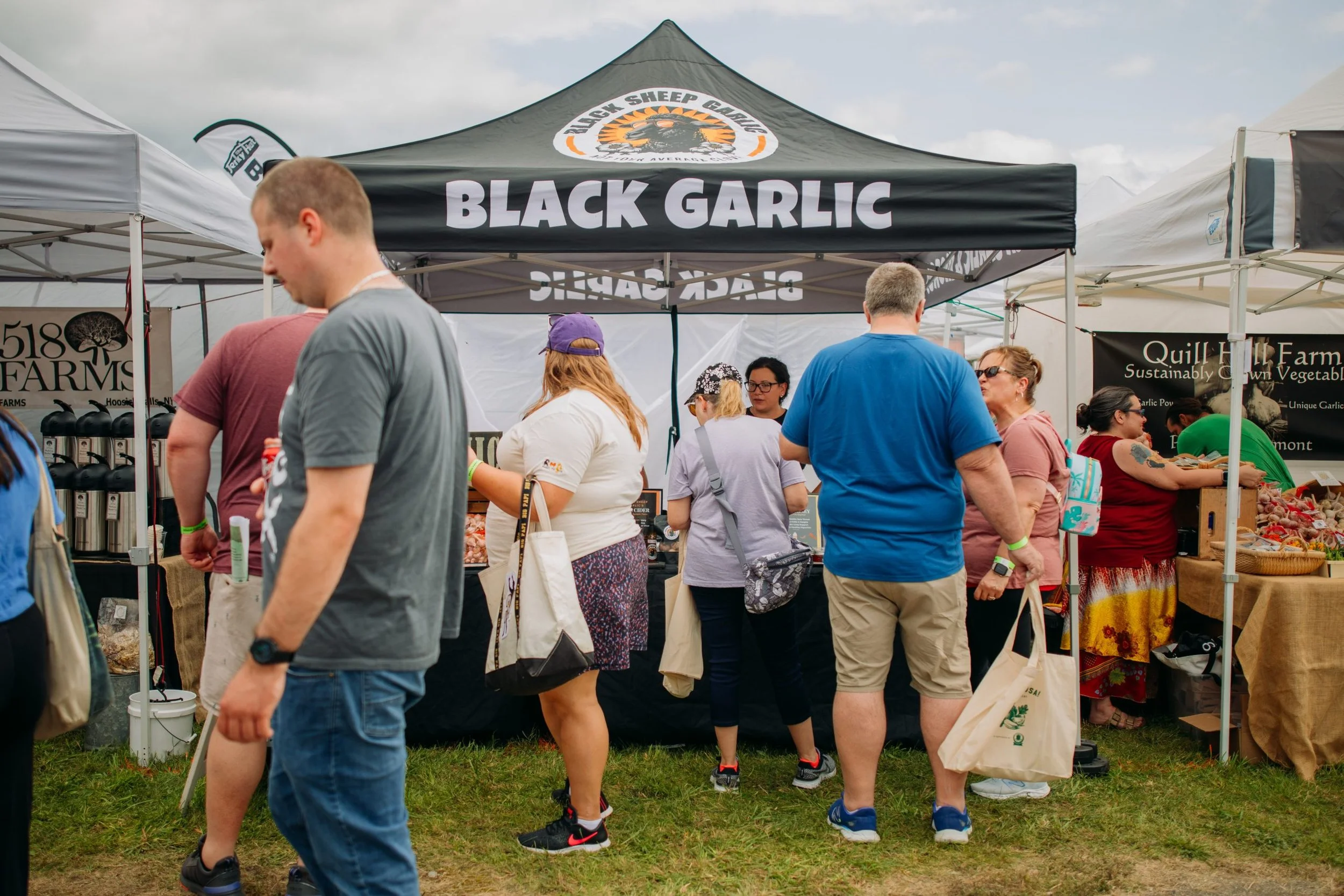 People at an outdoor farmers' market browsing and shopping at various stalls, with a black garlic vendor's tent. The tent has a logo with a sheep and the text 'Black Sheep Grown'