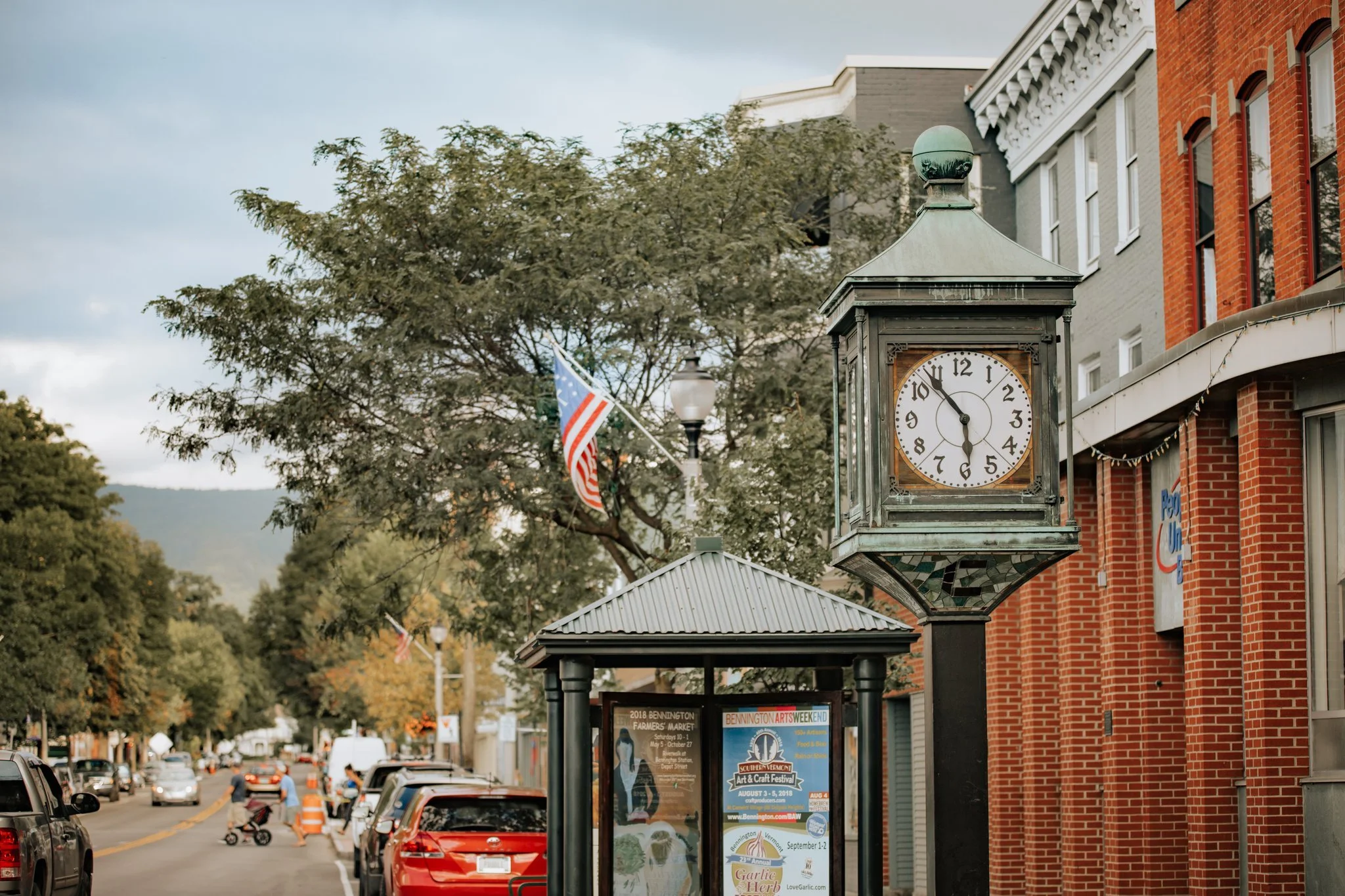 Street scene with a vintage clock tower, American flags, parked cars, trees, and buildings.