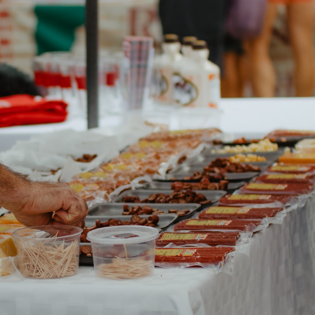Food vendor's table with various packaged meats, sauces, and toppings, including sliced sausages, chopped meats, and condiment bottles, at an outdoor event.