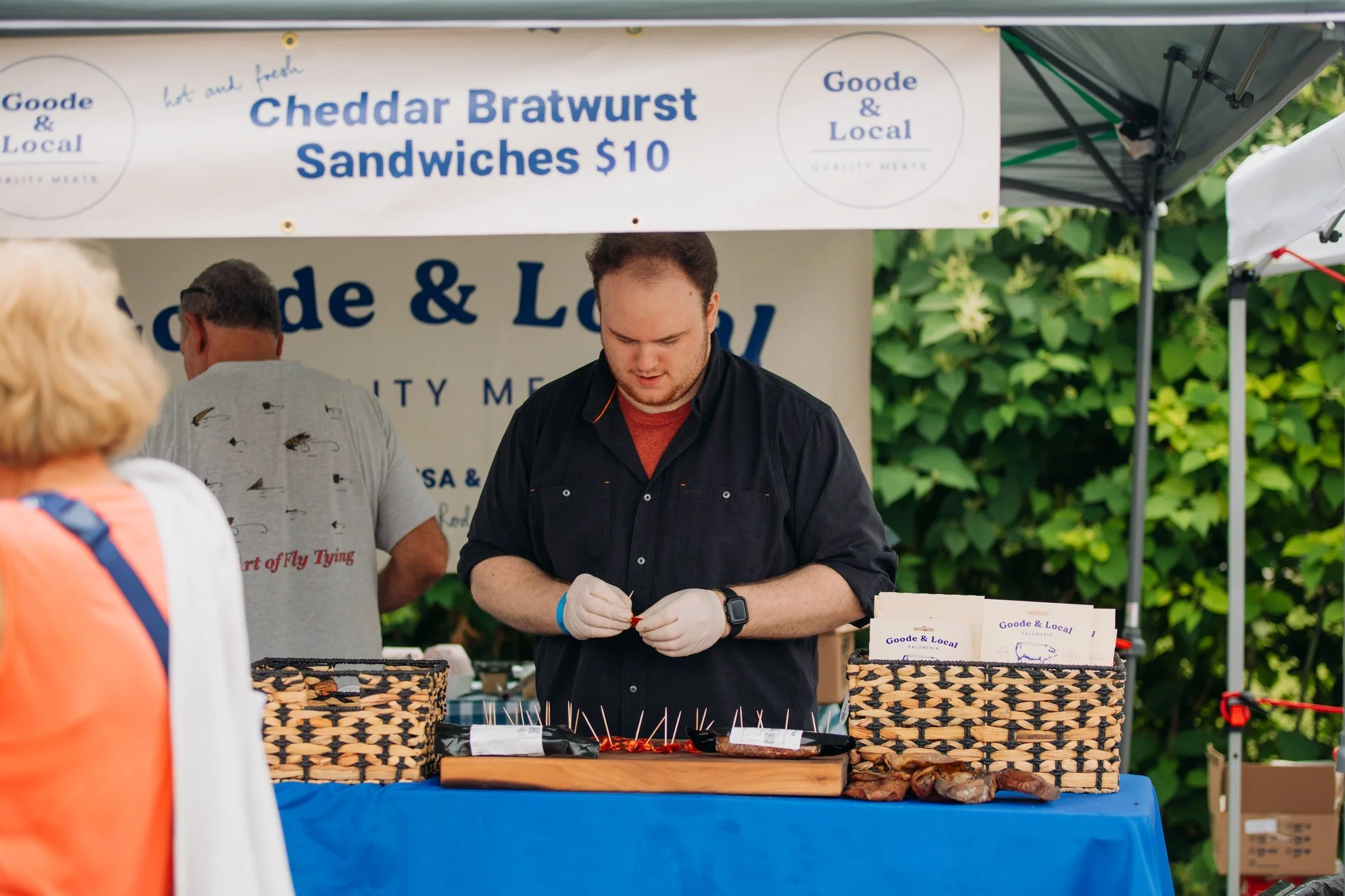 A man working at a food stall selling cheddar bratwurst sandwiches for $10 at an outdoor market, with other customers around and a sign in the background.