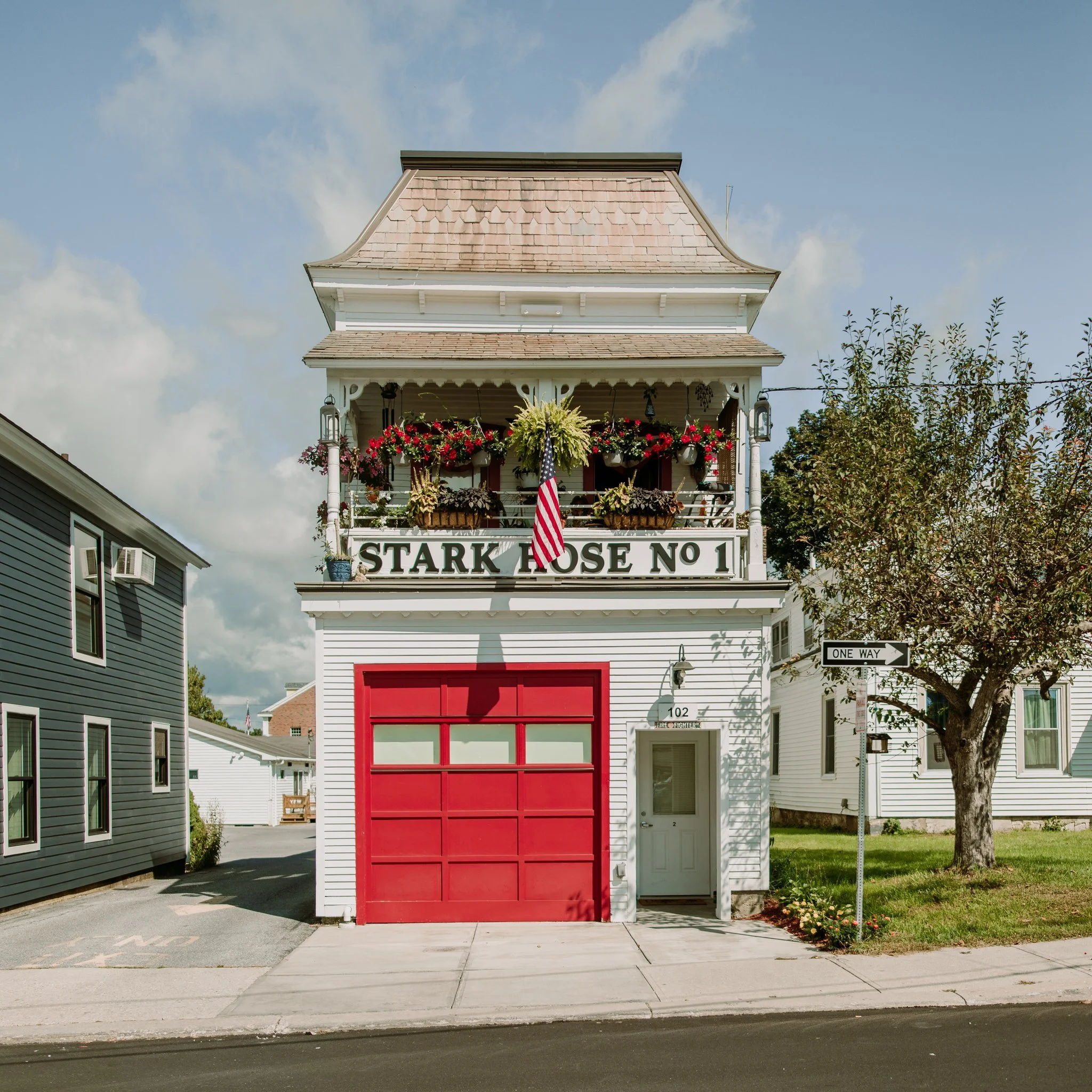 A white multi-story building with a red garage door and a white door next to it. The building has a sign that reads 'STARK ROSE NO 1' and a balcony with hanging plants and an American flag. To the right, a tree is visible, and there is a one-way stre