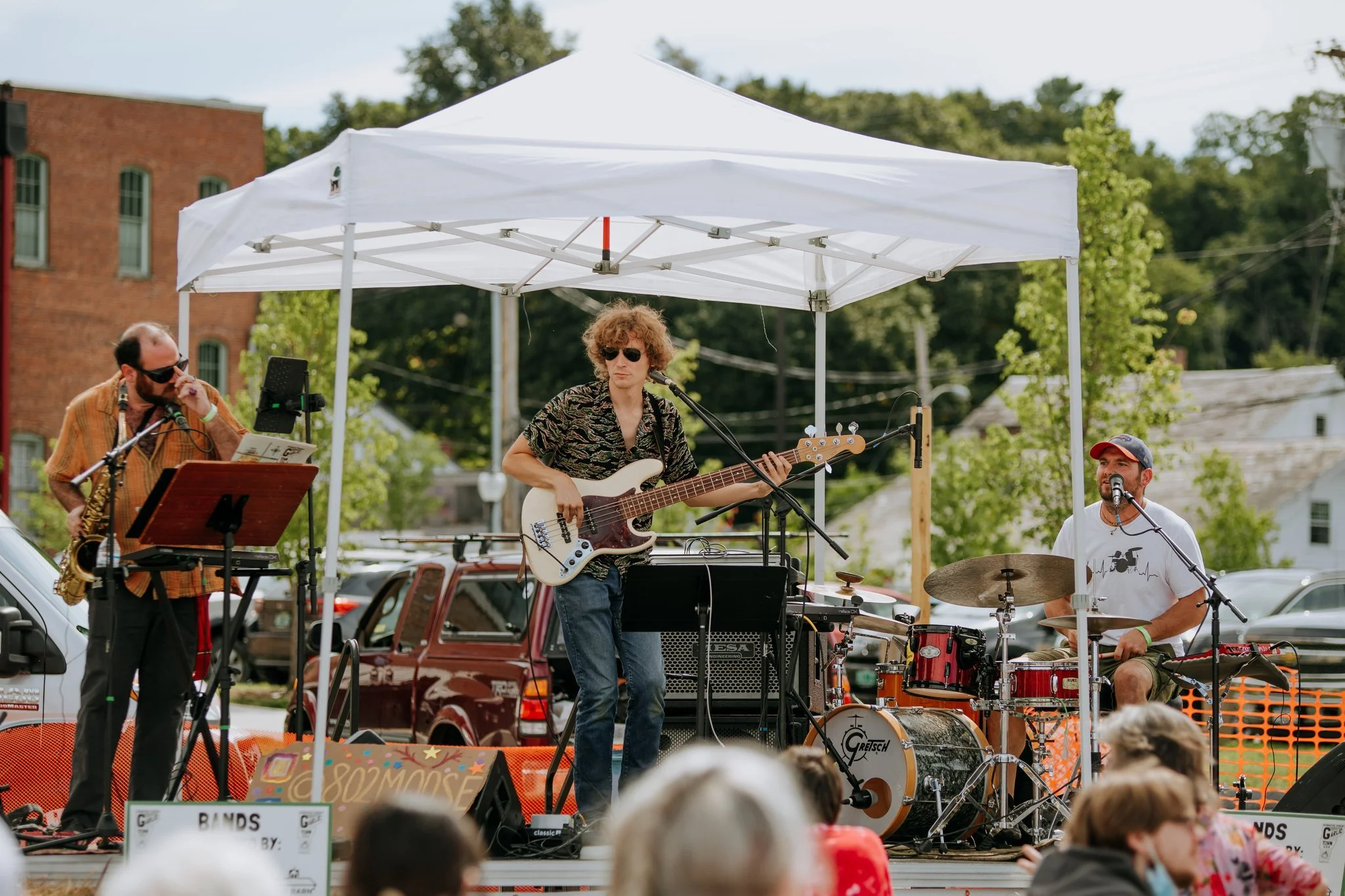 A band performs on an outdoor stage with three musicians: one on saxophone, one on bass guitar, and one on drums, under a white canopy tent. Audience members watch in the foreground.